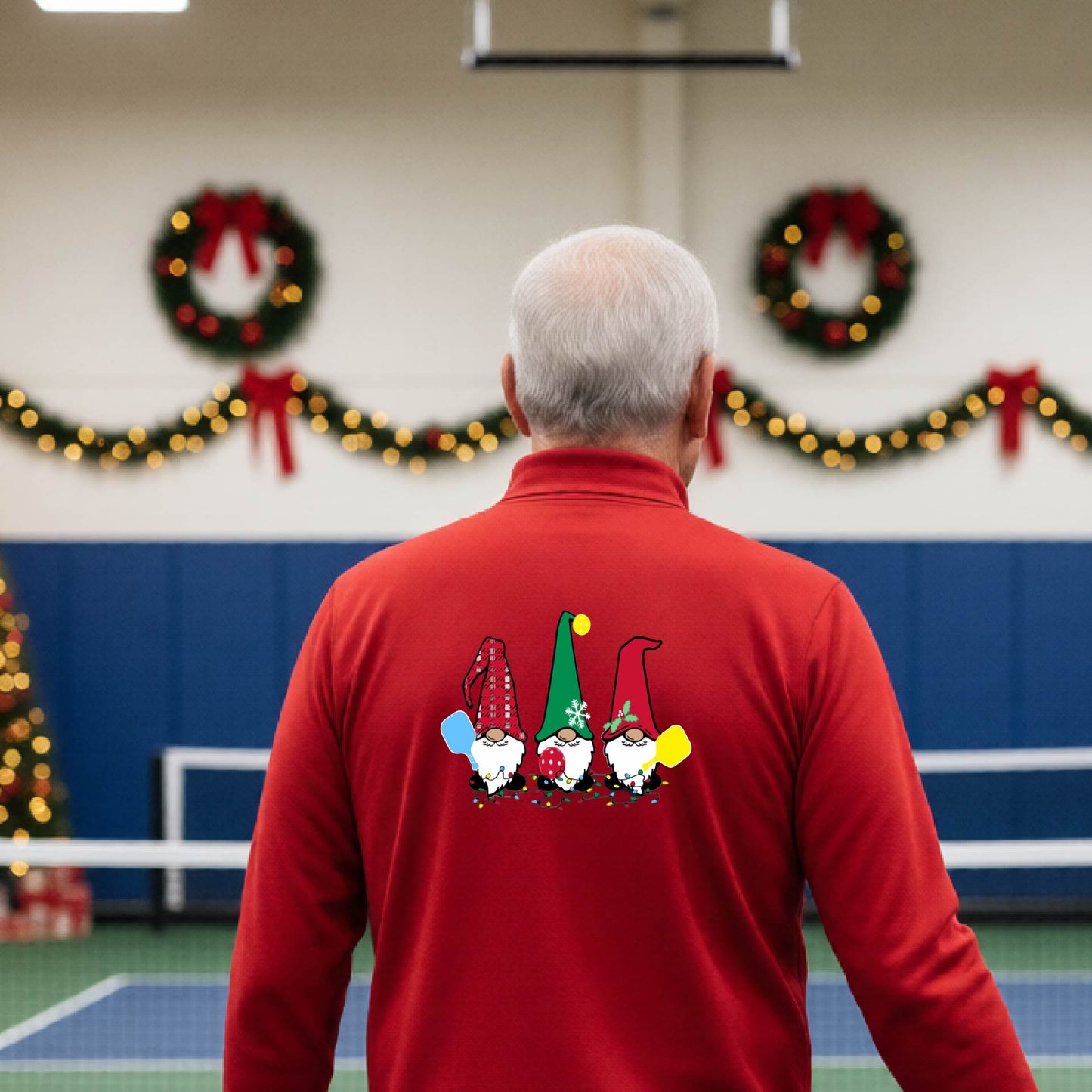 A man viewed from behind wearing a red long-sleeve quarter-zip pullover with a graphic of three Christmas gnomes on the back holding pickleball paddles, standing on pickleball court with holiday garlands in the background.