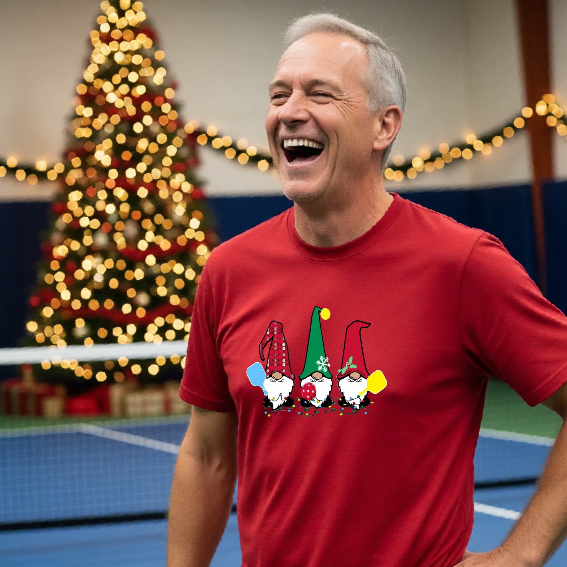 A man laughing on a pickleball court, wearing a red short-sleeve shirt with three colorful gnomes holding pickleball paddles graphic on the chest. The background features warm string lights and a decorated Christmas tree.
