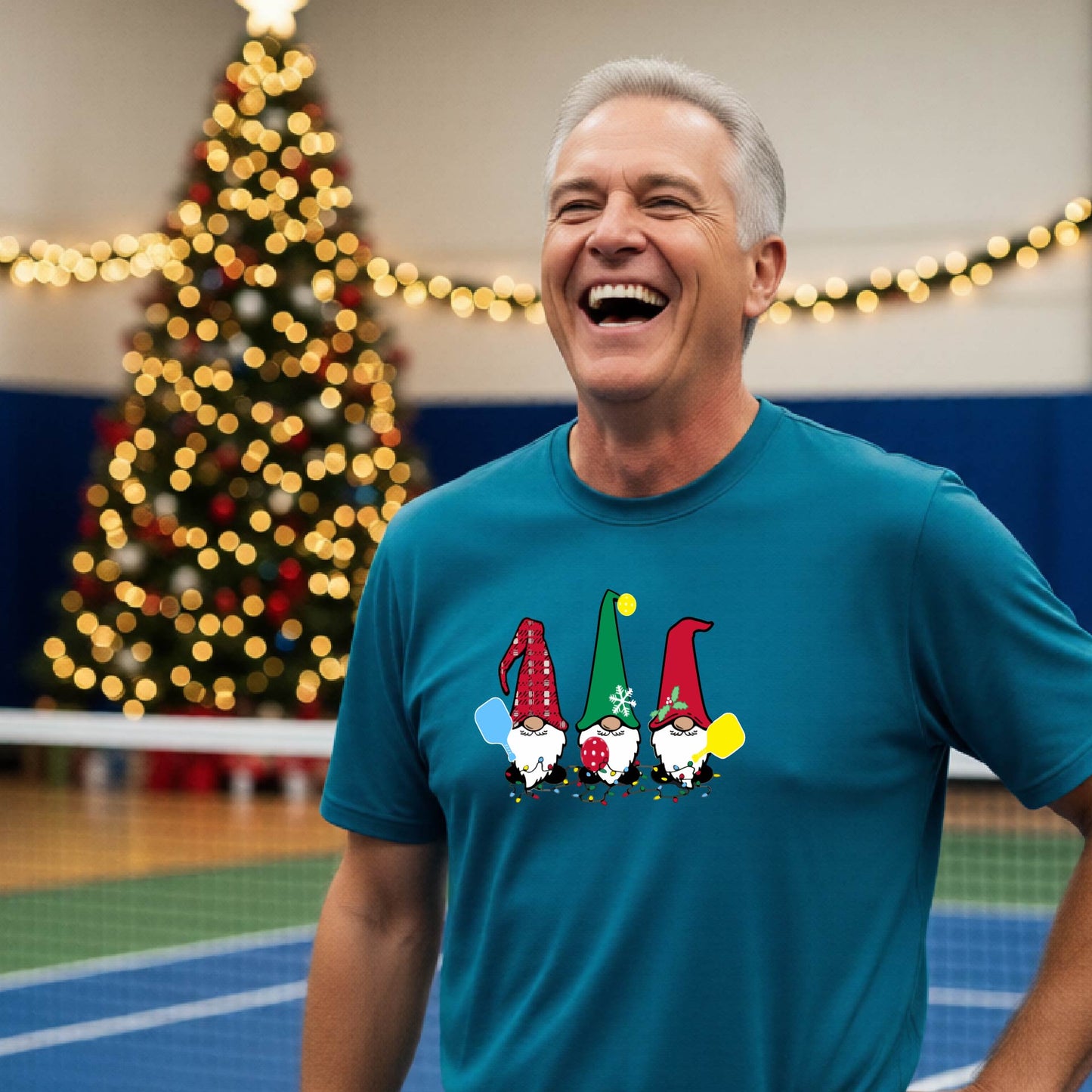 A man laughing on a pickleball court, wearing a blue short-sleeve shirt with three colorful gnomes holding pickleball paddles graphic on the chest. The background features warm string lights and a decorated Christmas tree.