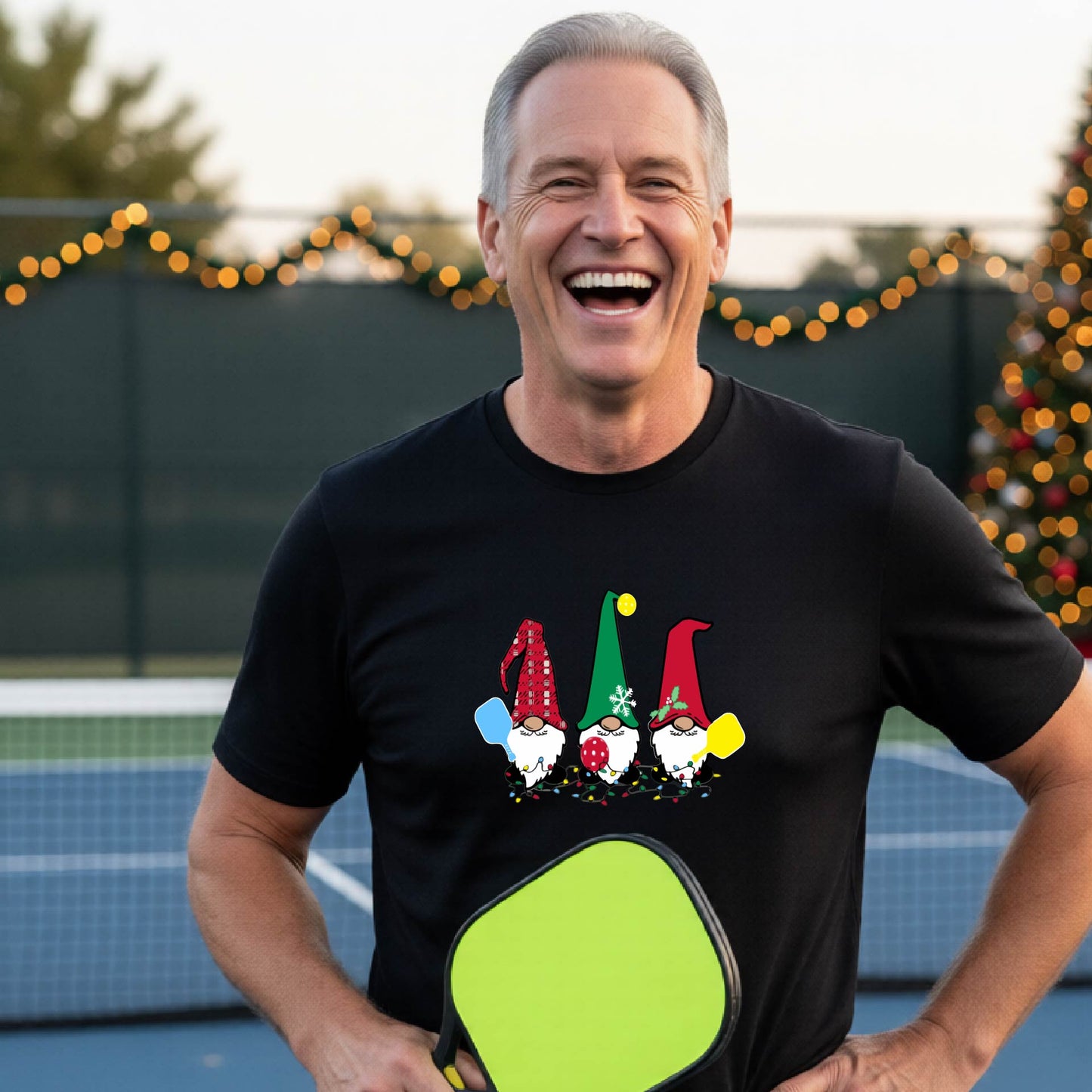 A man laughing on a pickleball court, wearing a black short-sleeve shirt with three colorful gnomes holding pickleball paddles graphic on the chest. The background features warm string lights and a decorated Christmas tree.