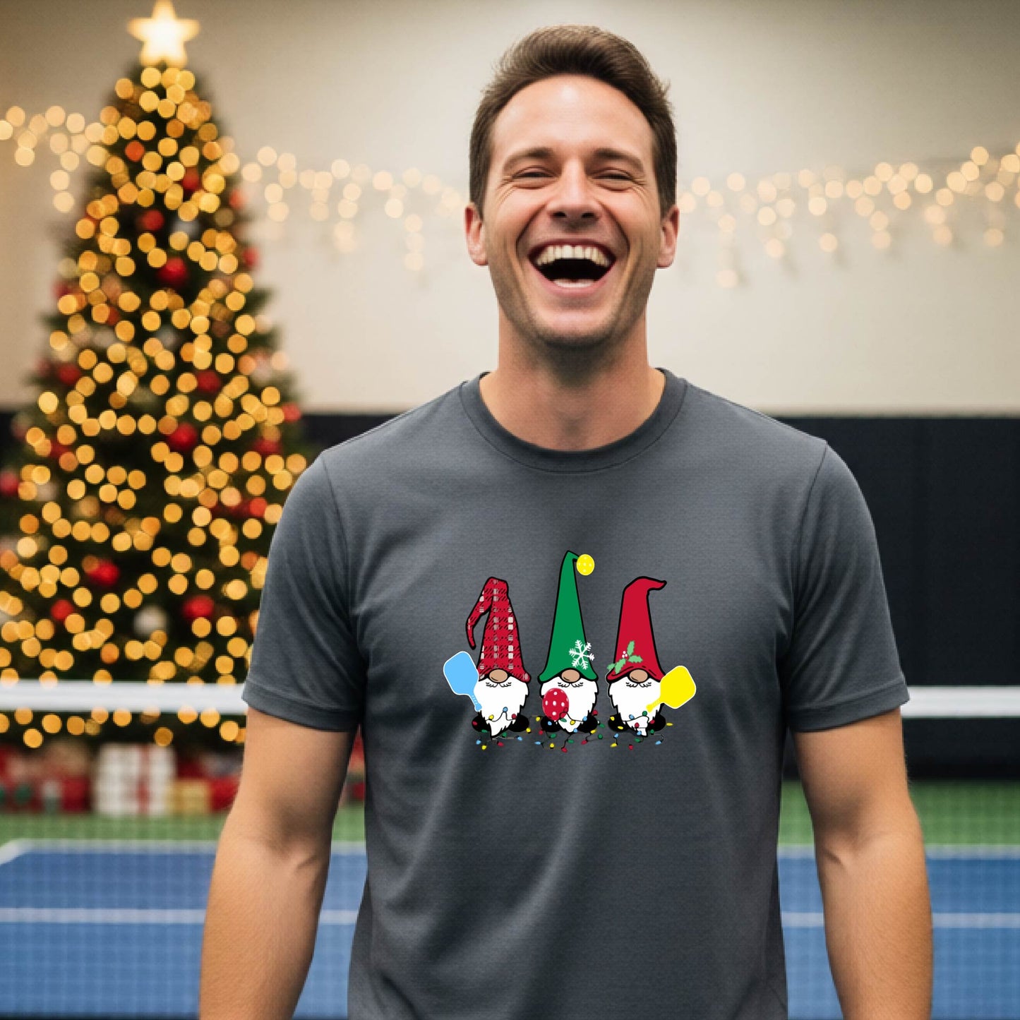 A man laughing on a pickleball court, wearing a gray short-sleeve shirt with three colorful gnomes holding pickleball paddles graphic on the chest. The background features warm string lights and a decorated Christmas tree.