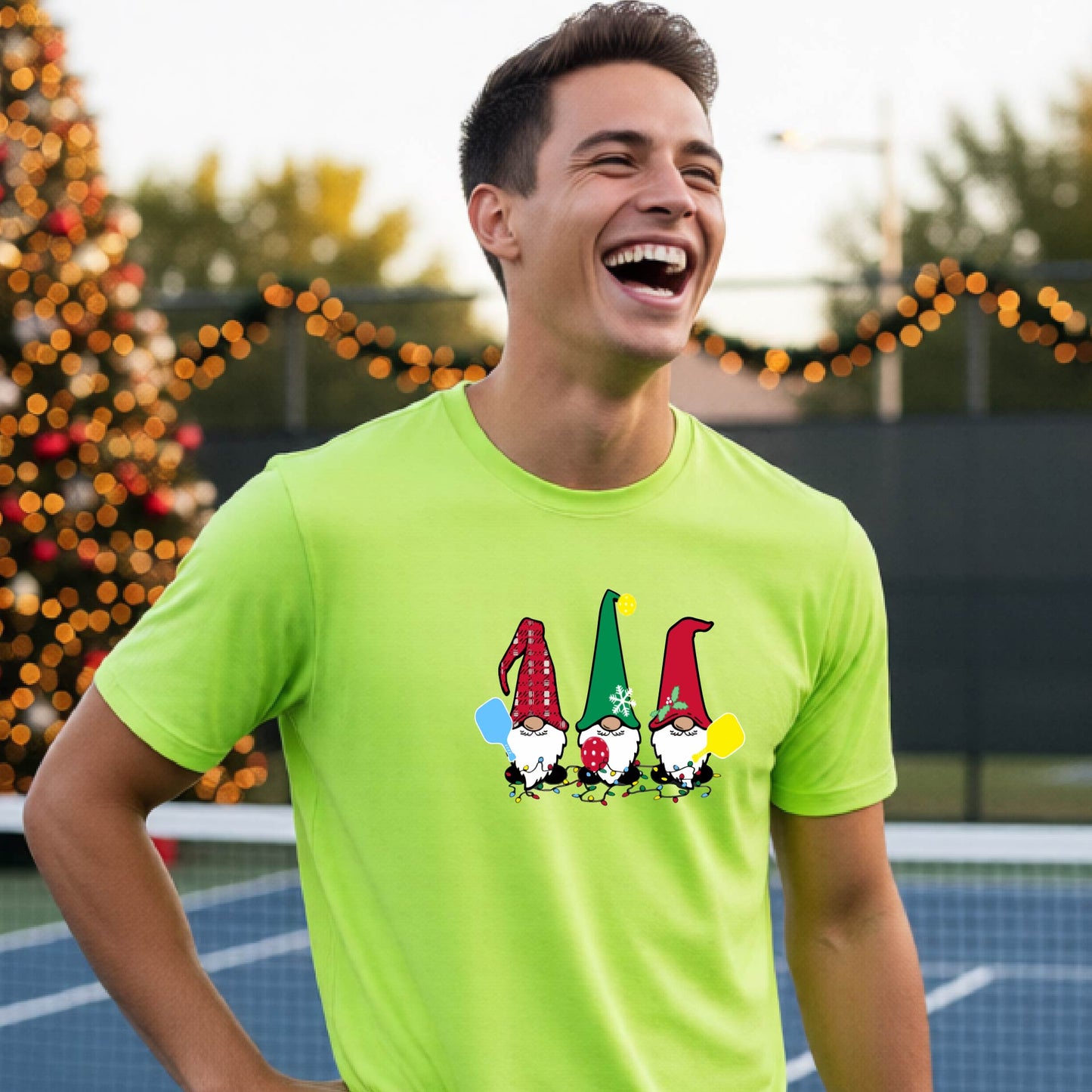 A man laughing on a pickleball court, wearing a lime green short-sleeve shirt with three colorful gnomes holding pickleball paddles graphic on the chest. The background features warm string lights and a decorated Christmas tree.