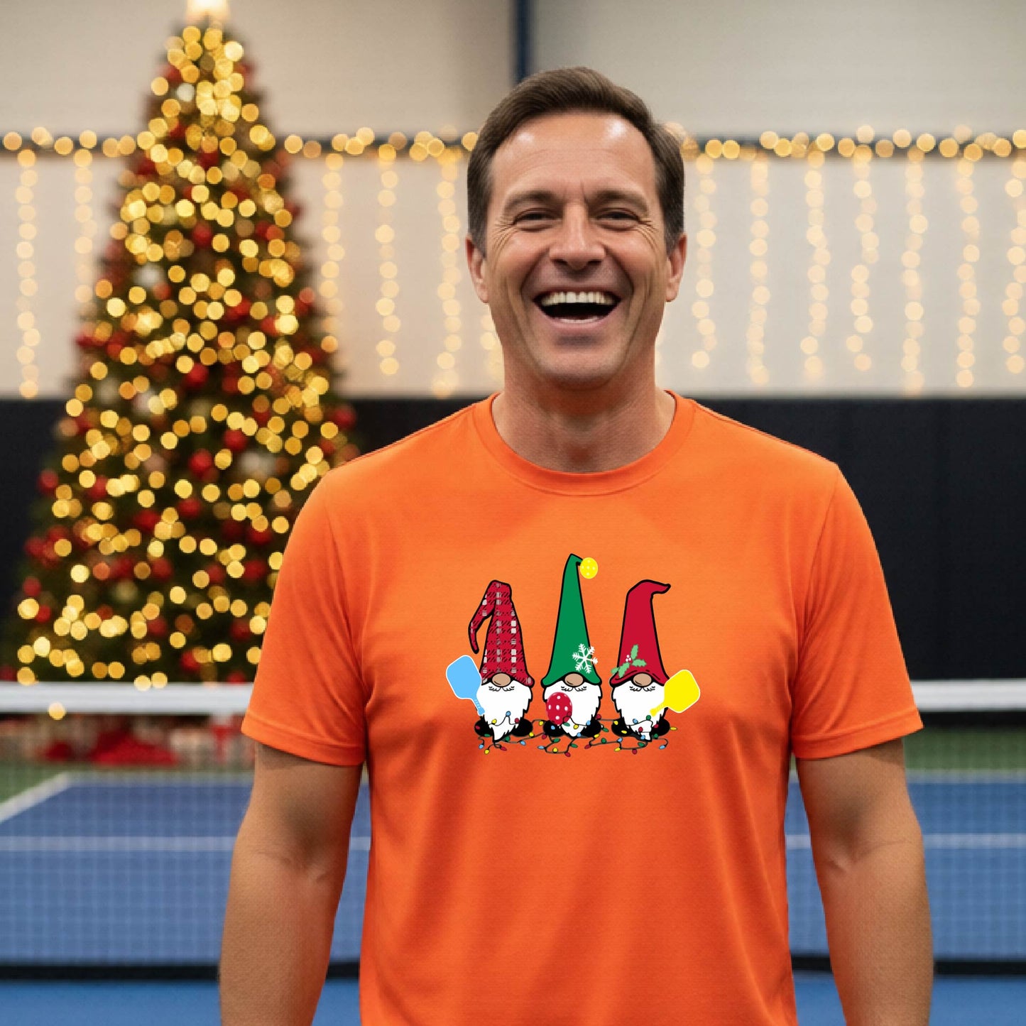 A man laughing on a pickleball court, wearing an orange short-sleeve shirt with three colorful gnomes holding pickleball paddles graphic on the chest. The background features warm string lights and a decorated Christmas tree.