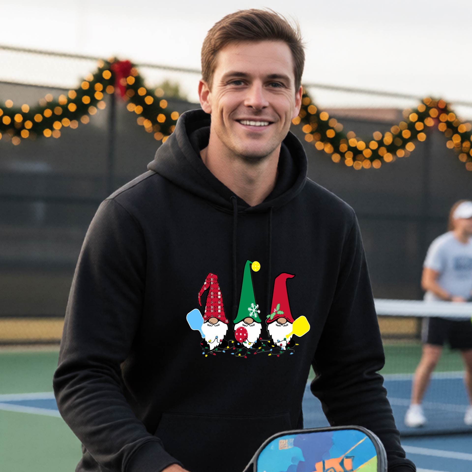 A smiling man wearing a black hoodie with a gnome graphic holding pickleball paddles stands on a picleball court. The hoodie features three colorful pickleball gnomes on the chest.
