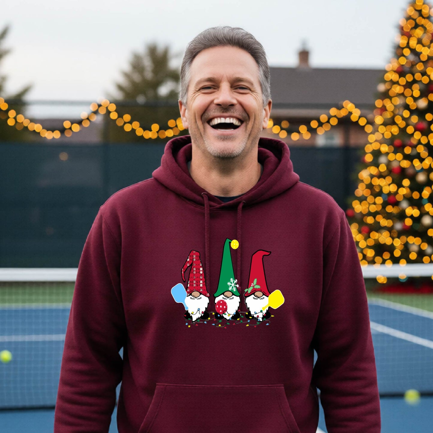 A smiling man wearing a maroon hoodie with a gnome graphic holding pickleball paddles stands on a picleball court. The hoodie features three colorful pickleball gnomes on the chest.
