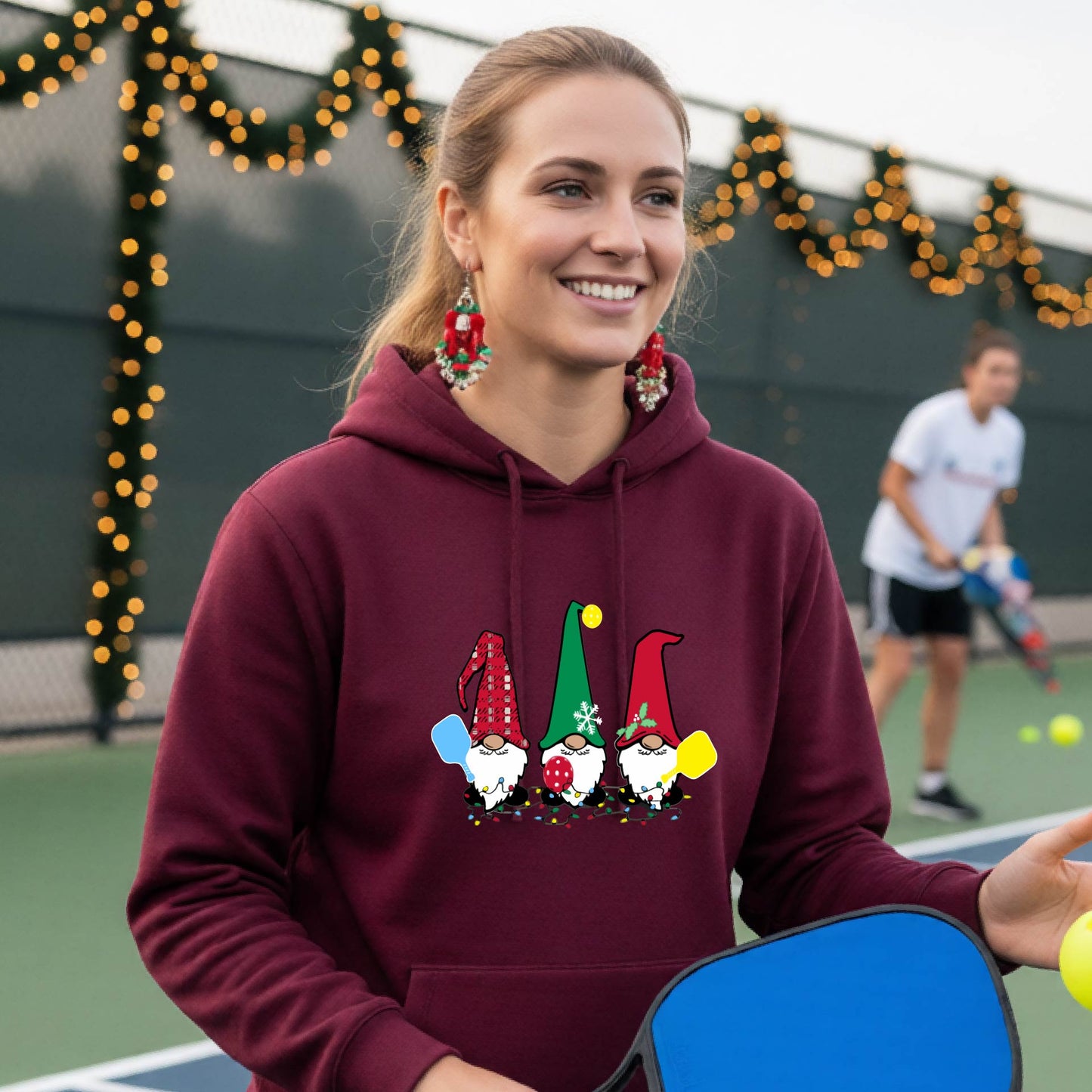 A smiling woman wearing a maroon hoodie with a gnome graphic holding pickleball paddles stands on a picleball court. The hoodie features three colorful pickleball gnomes on the chest.