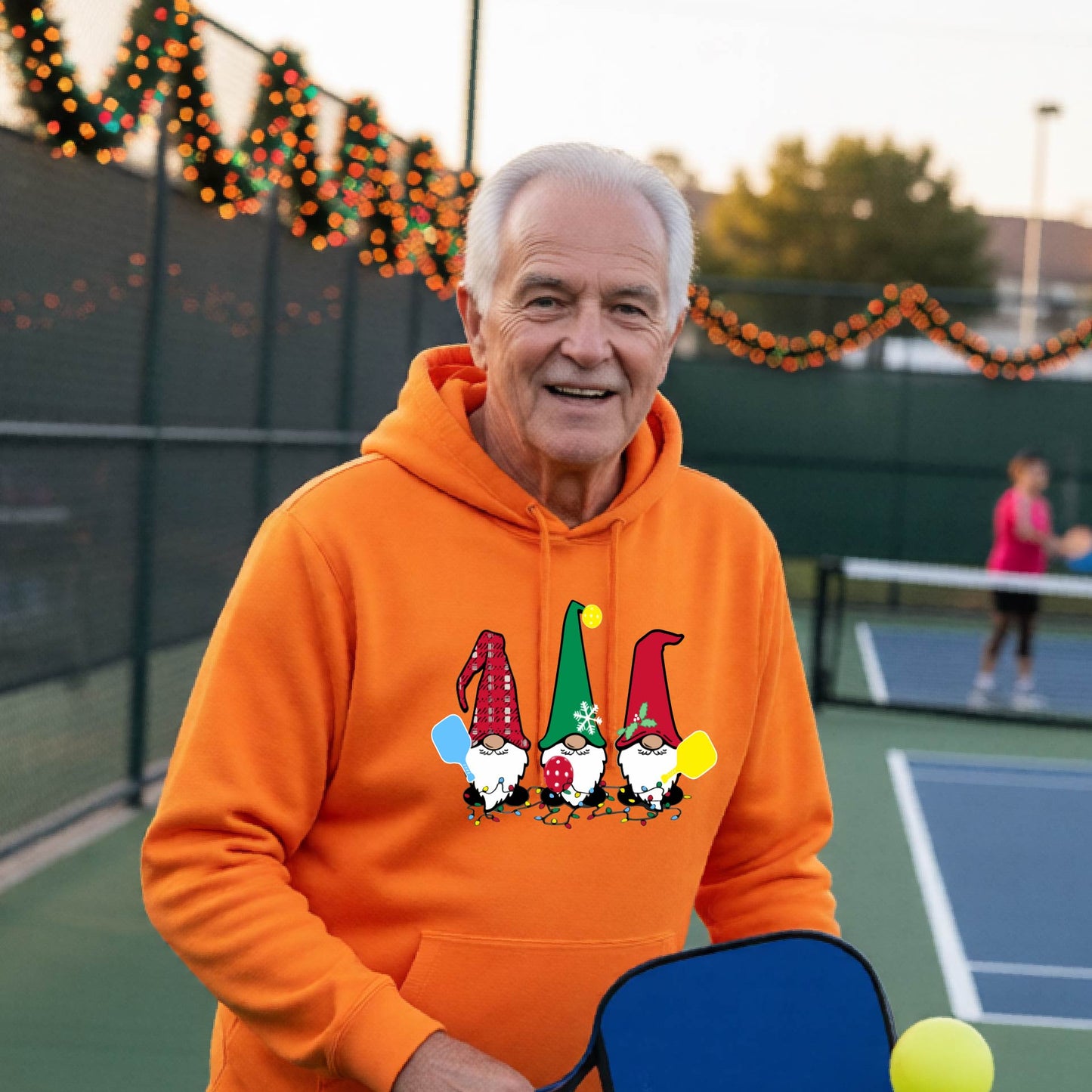 A smiling man wearing a bright orange hoodie with a gnome graphic holding pickleball paddles stands on a picleball court. The hoodie features three colorful pickleball gnomes on the chest.