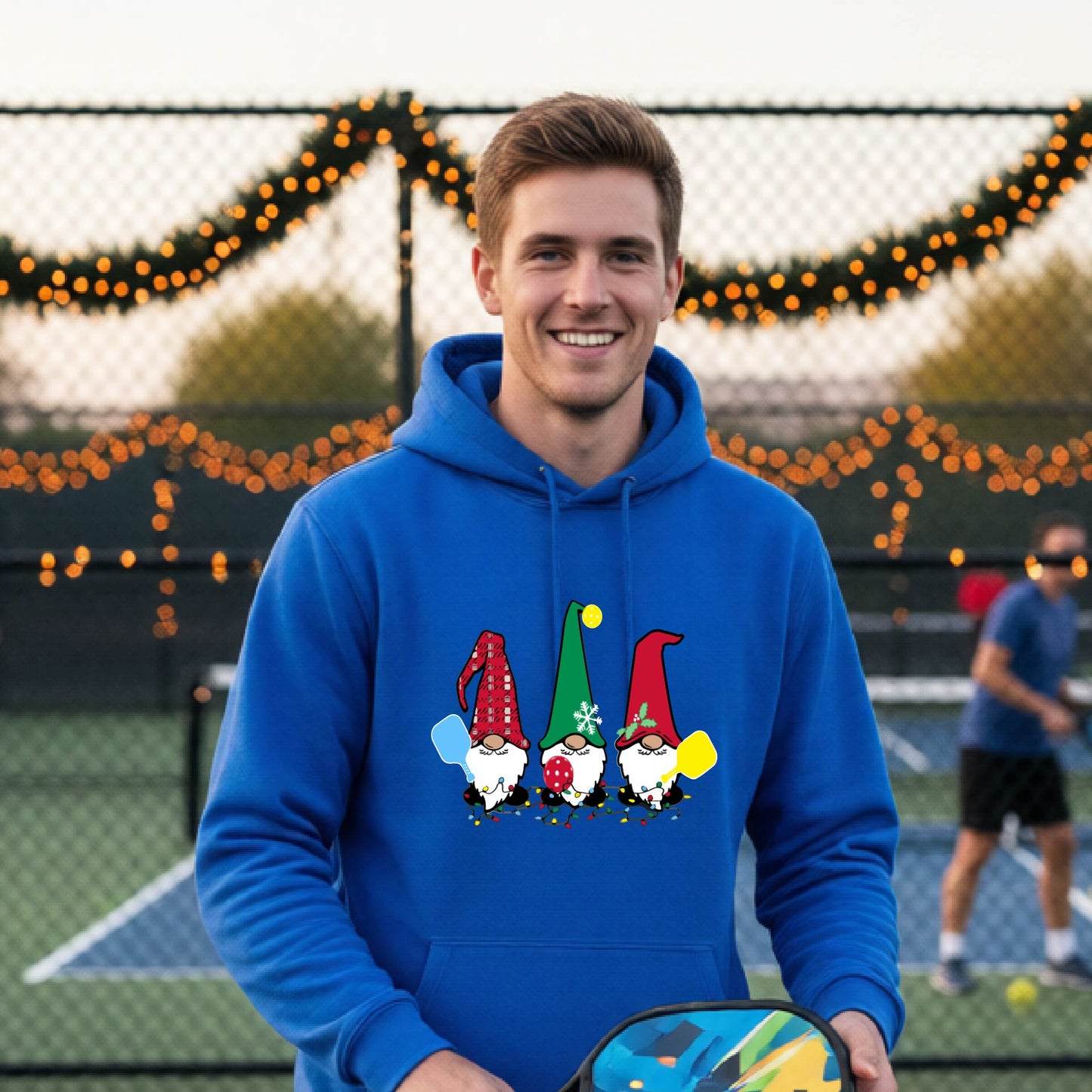 A smiling man wearing a bright royal blue hoodie with a gnome graphic holding pickleball paddles stands on a pickleball court. The hoodie features three colorful pickleball gnomes on the chest.