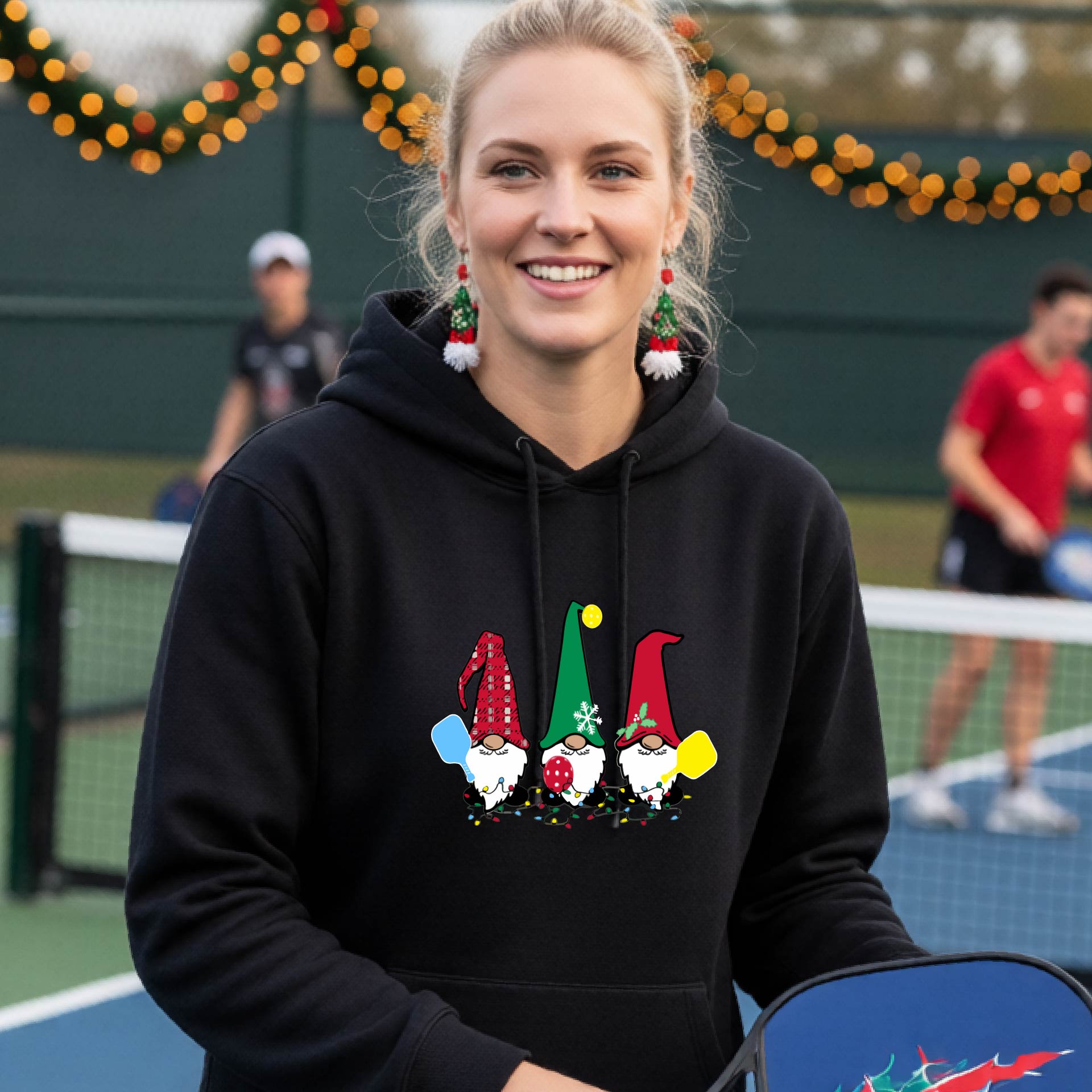 A smiling woman wearing a bright green hoodie with a gnome graphic holding pickleball paddles stands on a pickleball court. The hoodie features three colorful pickleball gnomes on the chest.