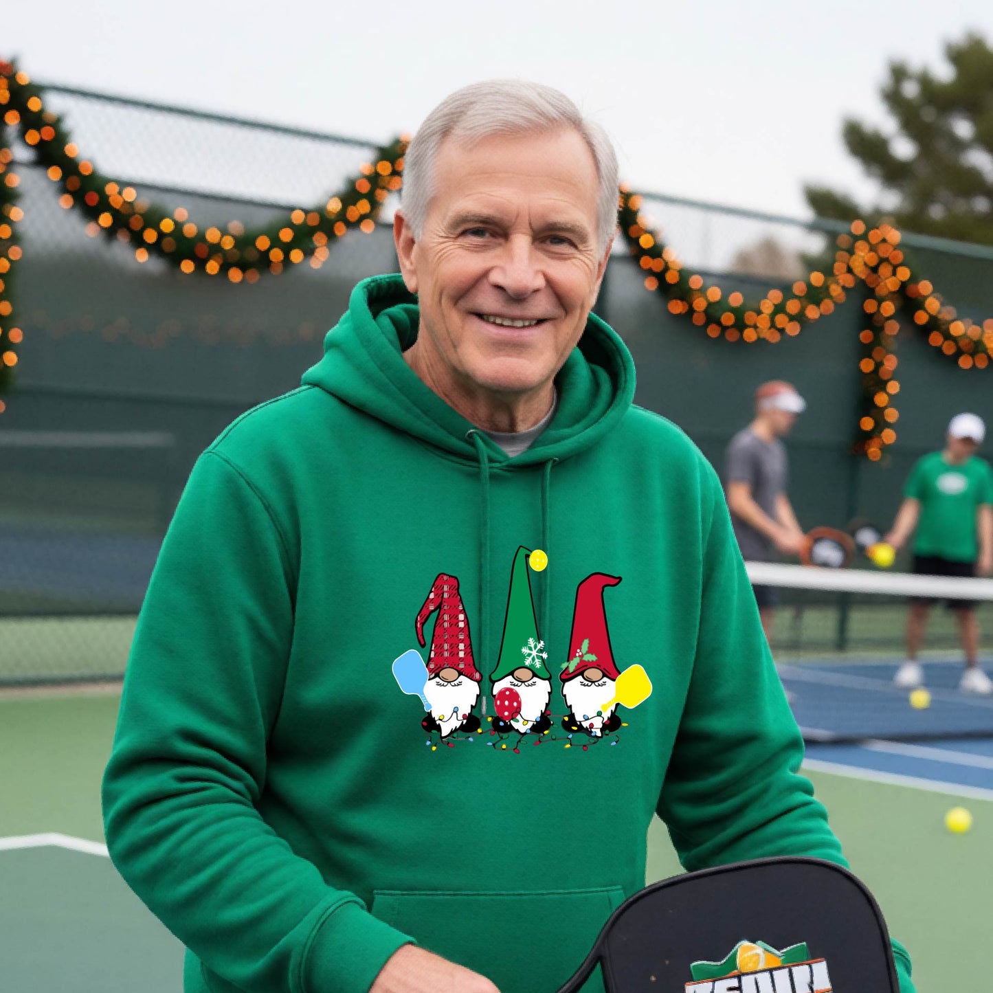 A smiling man wearing a bright green hoodie with a gnome graphic holding pickleball paddles stands on a picleball court. The hoodie features three colorful pickleball gnomes on the chest.
