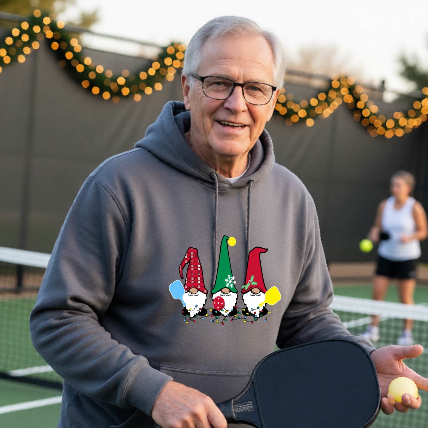 A smiling man wearing a gray hoodie with a gnome graphic holding pickleball paddles stands on a picleball court. The hoodie features three colorful pickleball gnomes on the chest.