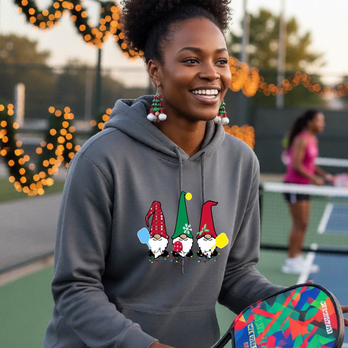 A smiling woman wearing a gray hoodie with a gnome graphic holding pickleball paddles stands on a picleball court. The hoodie features three colorful pickleball gnomes on the chest.