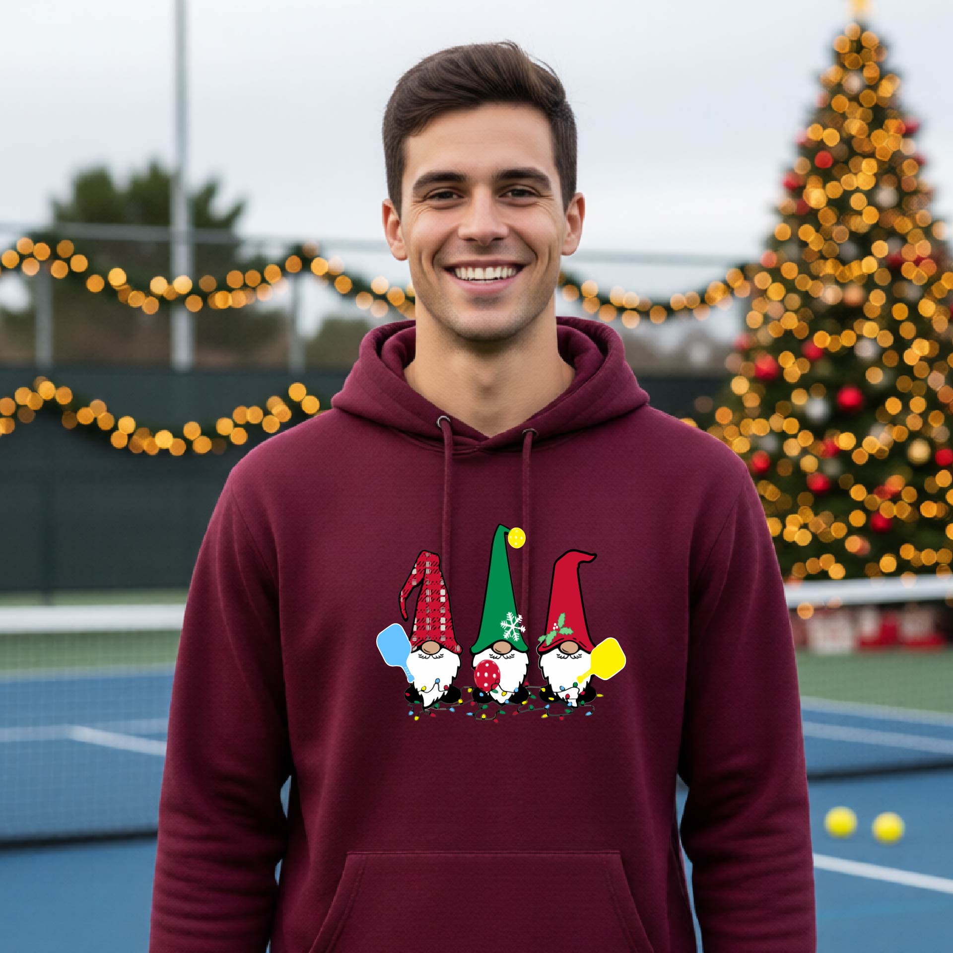 A smiling man wearing a maroon hoodie with a gnome graphic holding pickleball paddles stands on a picleball court. The hoodie features three colorful pickleball gnomes on the chest.