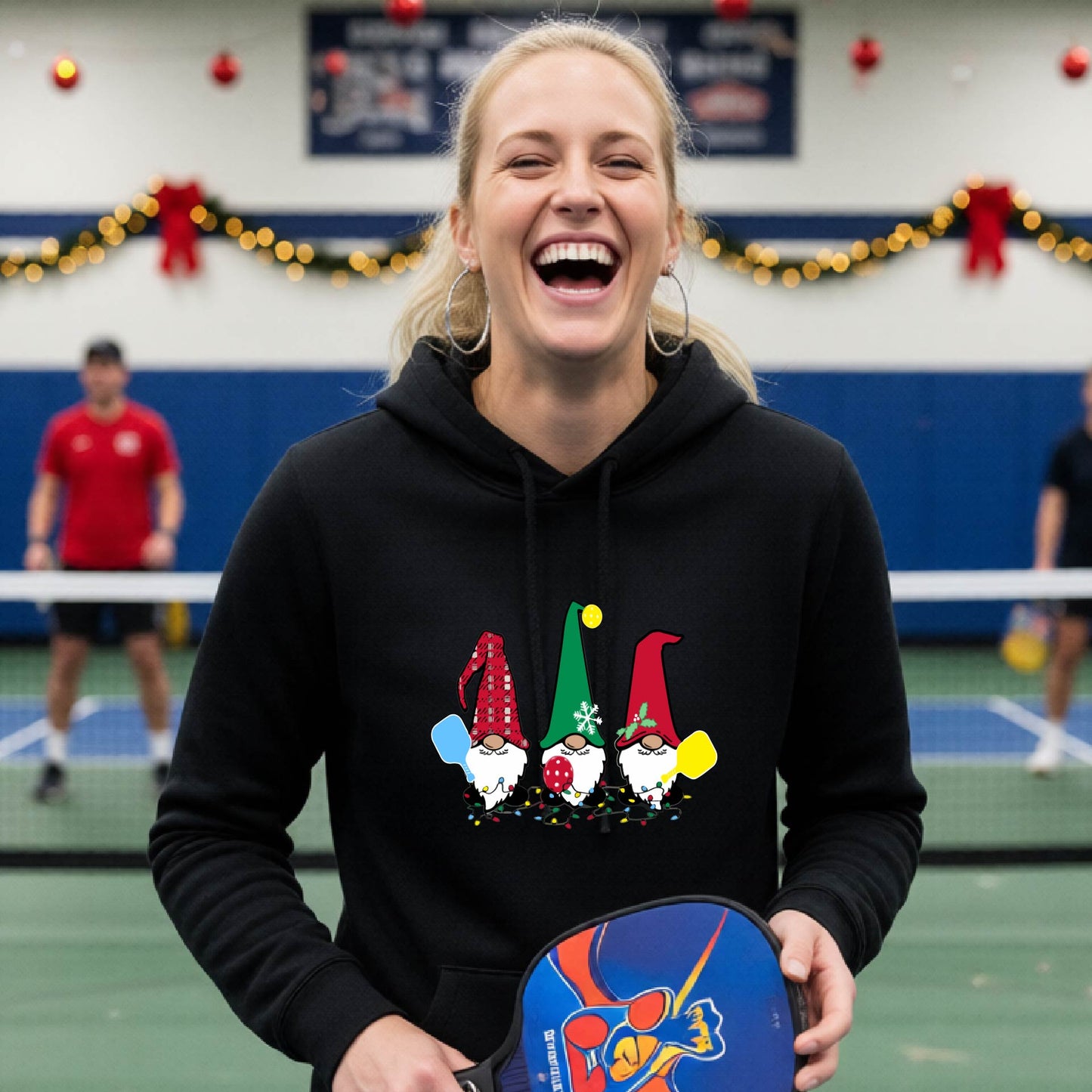 A black hooded sweatshirt featuring a pickleball graphic with gnomes wearing Chrsitmas hats and holding pickleball paddles and a Christmas themed bakground. 