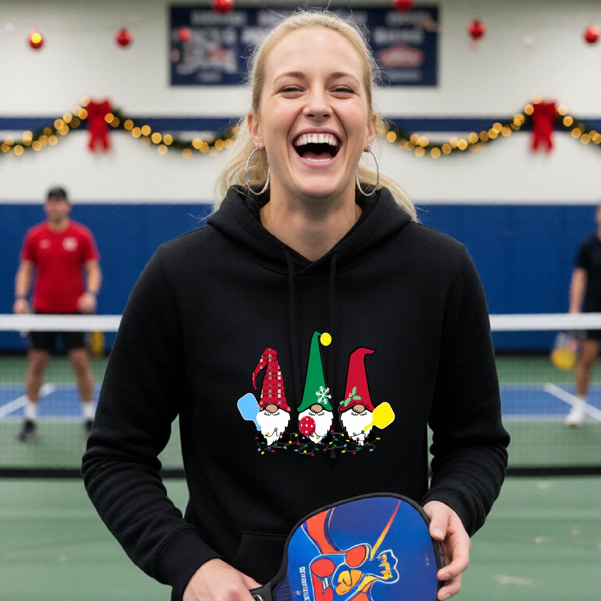 A black hooded sweatshirt featuring a pickleball graphic with gnomes wearing Chrsitmas hats and holding pickleball paddles and a Christmas themed bakground. 