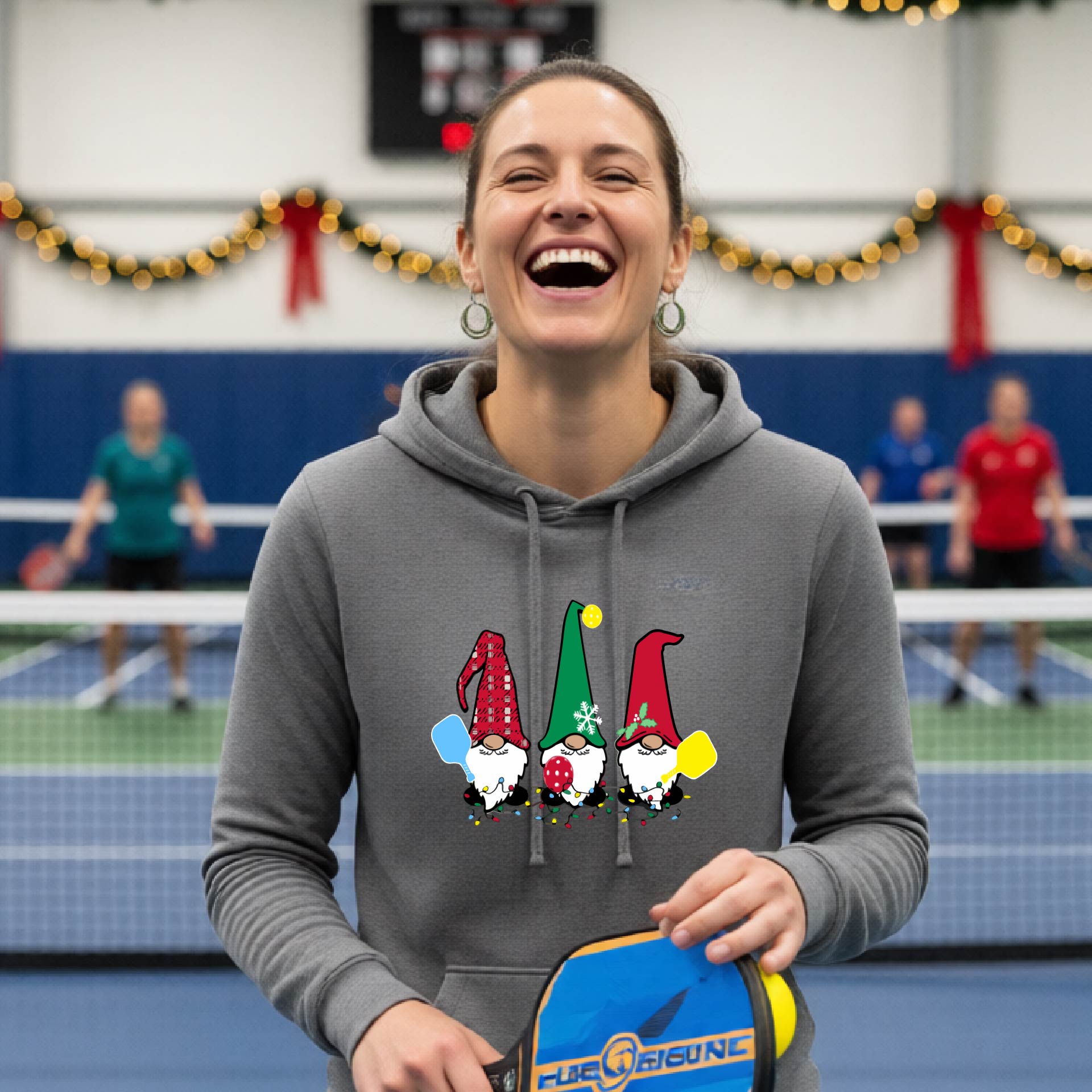 A gray hooded sweatshirt featuring a pickleball graphic with gnomes wearing Chrsitmas hats and holding pickleball paddles and a Christmas themed bakground. 