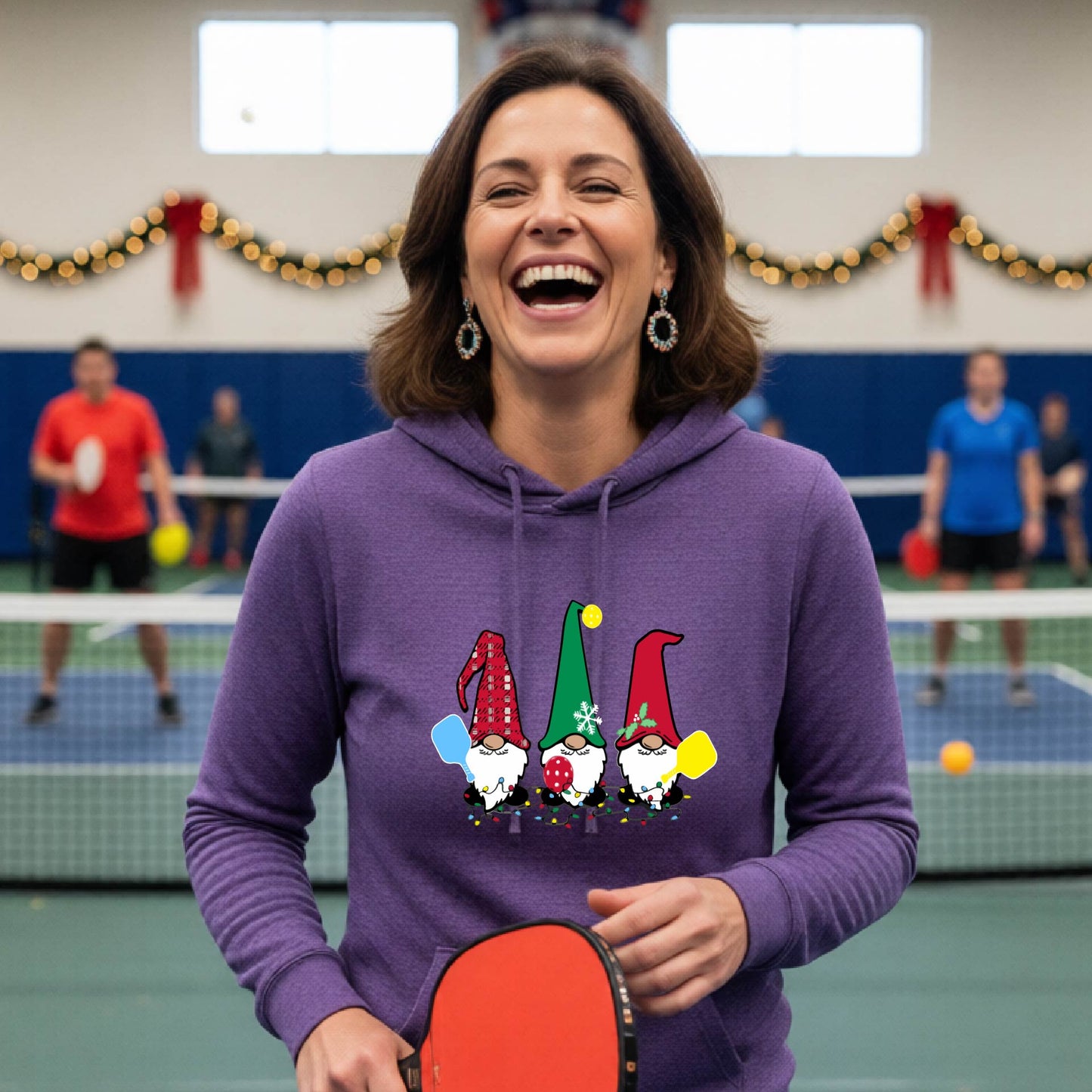 A purple hooded sweatshirt featuring a pickleball graphic with gnomes wearing Chrsitmas hats and holding pickleball paddles and a Christmas themed bakground. 