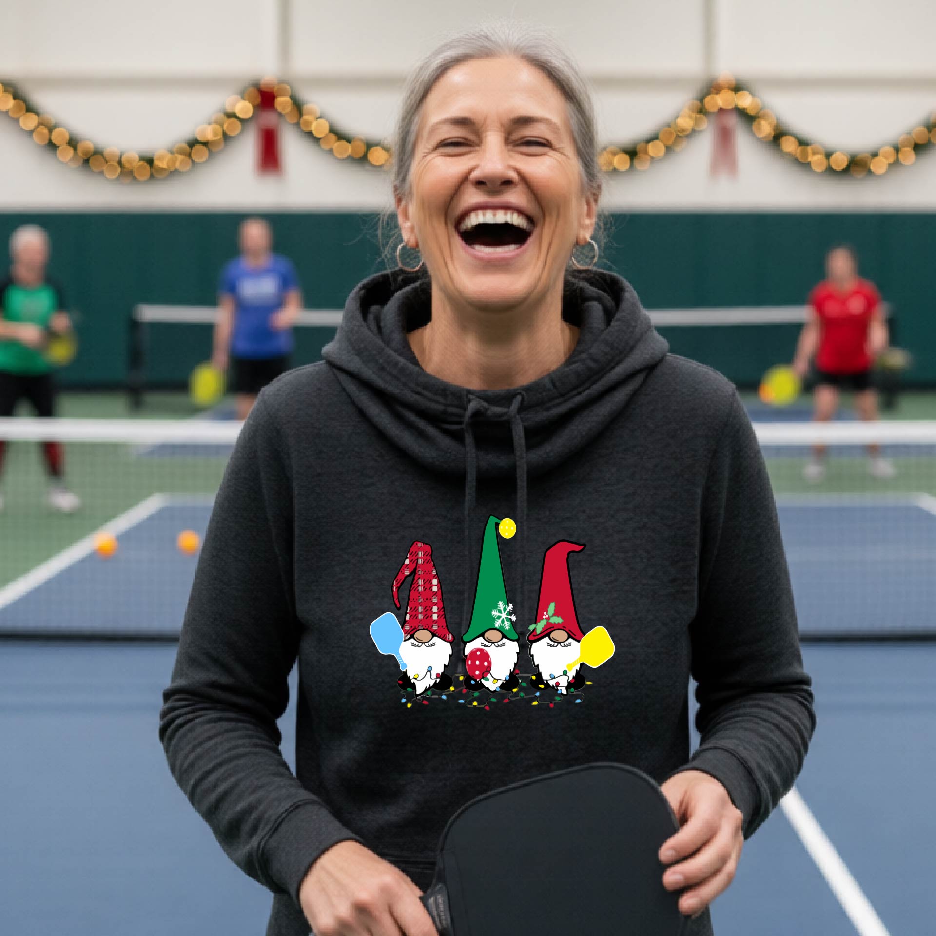 A black cowl-neck  hooded sweatshirt featuring a pickleball graphic with gnomes wearing Christmas hats and holding pickleball paddles and a Christmas themed background. 