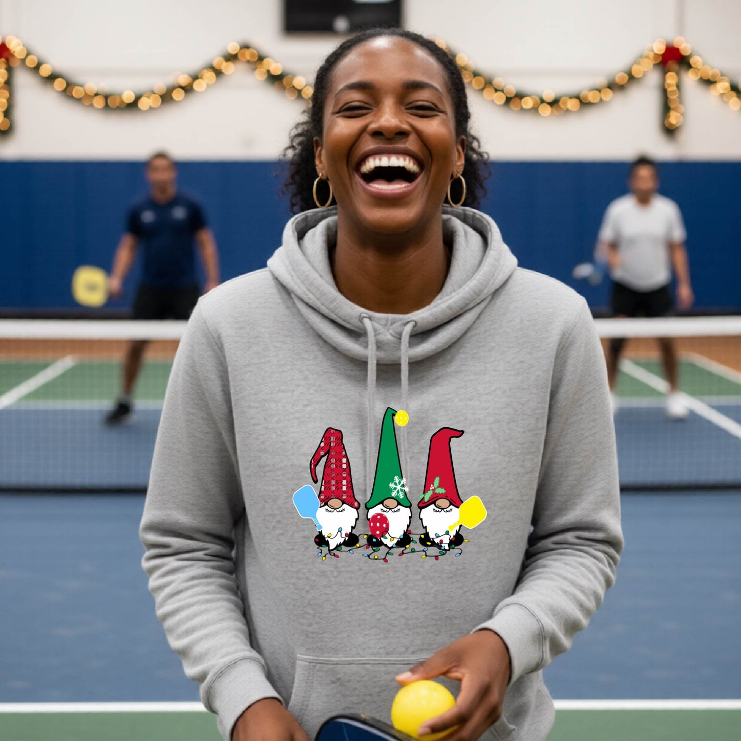 A grey cowl-neck  hooded sweatshirt featuring a pickleball graphic with gnomes wearing Christmas hats and holding pickleball paddles and a Christmas themed background. 