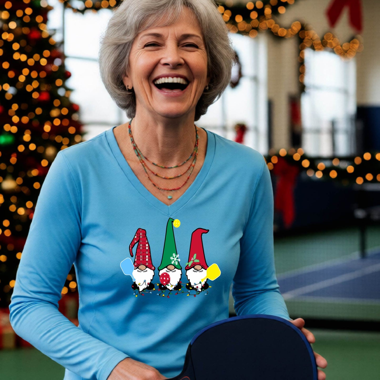 A woman laughing on a pickleball court, wearing a bright blue long-sleeve t-shirt with three colorful gnome holding pickleball paddles graphic on the chest. The background features warm string lights and a decorated Christmas tree.