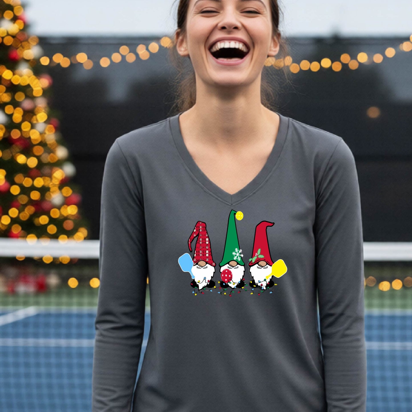 A woman laughing on a pickleball court, wearing a dark grey long-sleeve t-shirt with three colorful gnome holding pickleball paddles graphic on the chest. The background features warm string lights and a decorated Christmas tree.