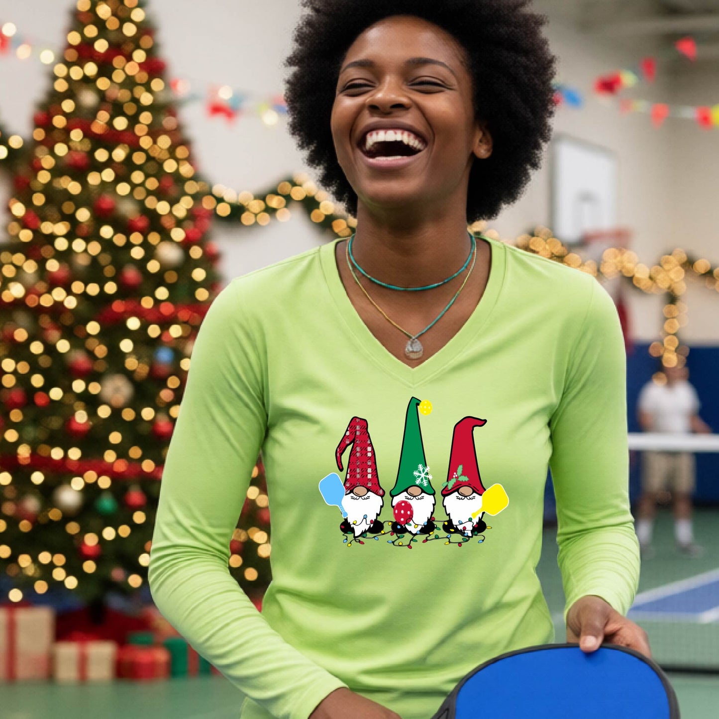 A woman laughing on a pickleball court, wearing a lime green long-sleeve t-shirt with three colorful gnome holding pickleball paddles graphic on the chest. The background features warm string lights and a decorated Christmas tree.