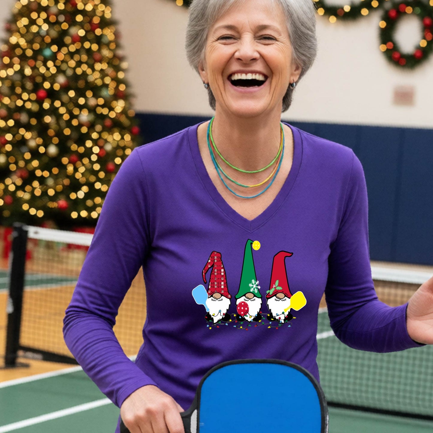 A woman laughing on a pickleball court, wearing a purple long-sleeve t-shirt with three colorful gnome holding pickleball paddles graphic on the chest. The background features warm string lights and a decorated Christmas tree. 