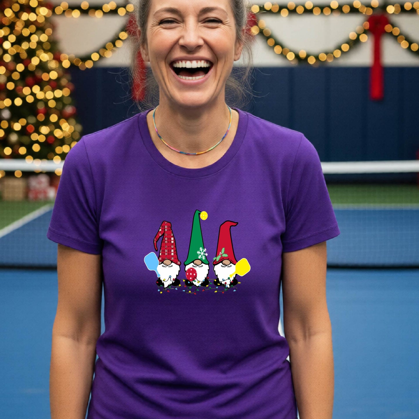 A smiling woman wearing a purple graphic t-shirt with three cartoon Christmas pickleball gnomes, standing in a gymnasium decorated with holiday lights and a Christmas tree.