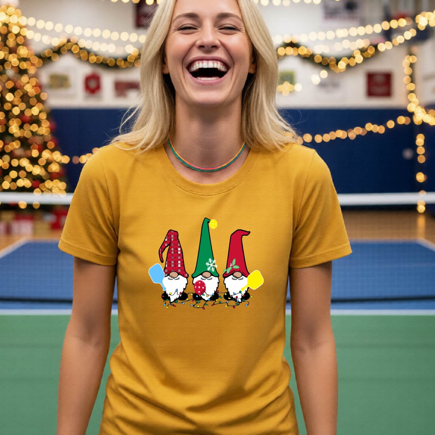 A smiling woman wearing a mustard yellow graphic t-shirt with three cartoon Christmas pickleball gnomes, standing in a gymnasium decorated with holiday lights and a Christmas tree.