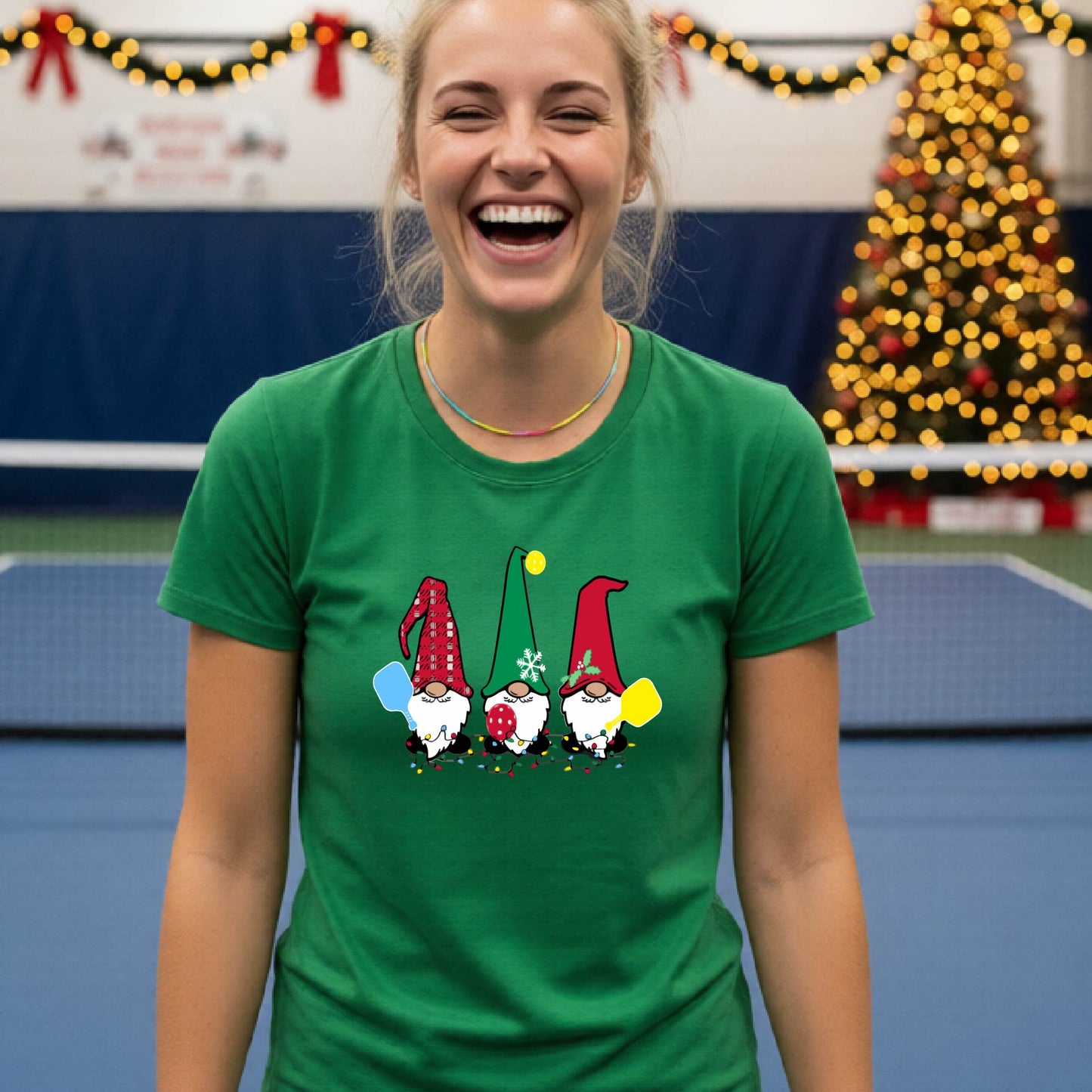 A smiling woman wearing a green graphic t-shirt with three cartoon Christmas pickleball gnomes, standing in a gymnasium decorated with holiday lights and a Christmas tree.