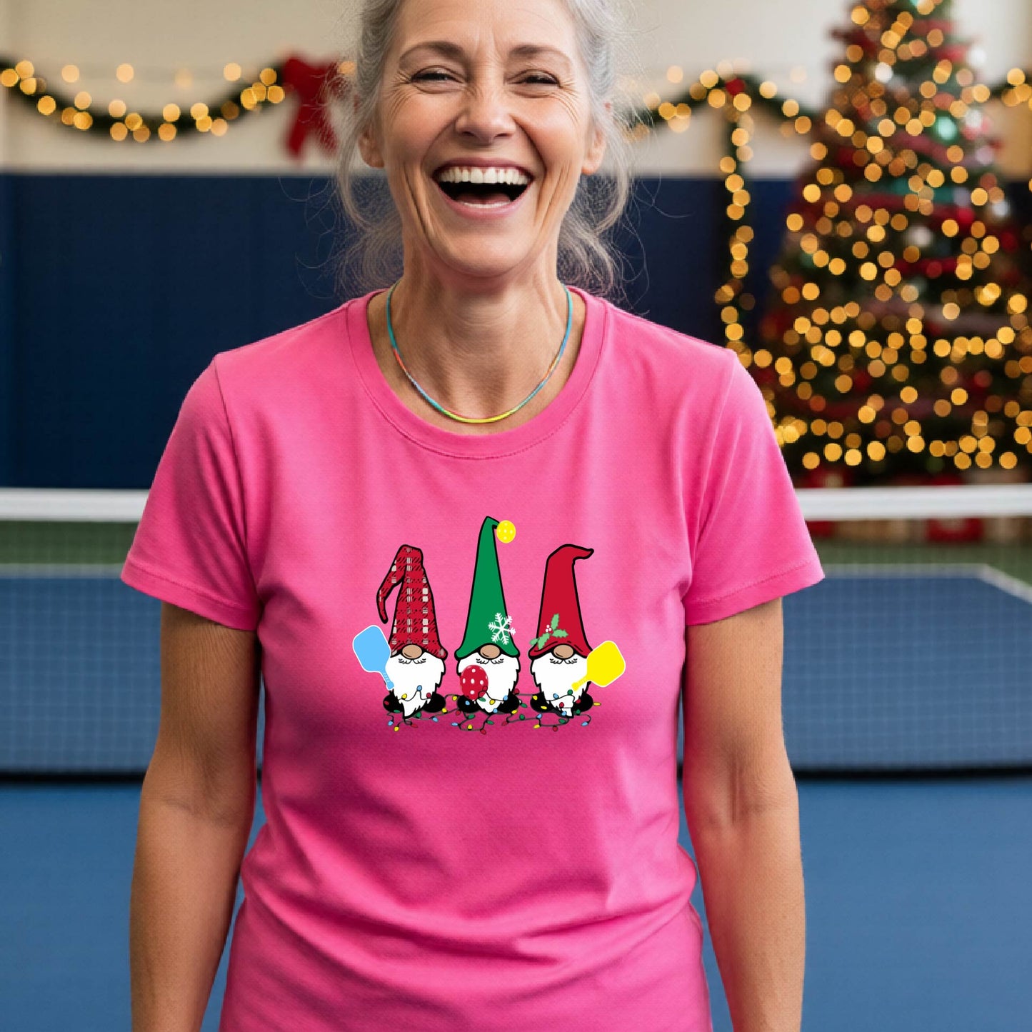 A smiling woman wearing a bright pink graphic t-shirt with three cartoon Christmas pickleball gnomes, standing in a gymnasium decorated with holiday lights and a Christmas tree.