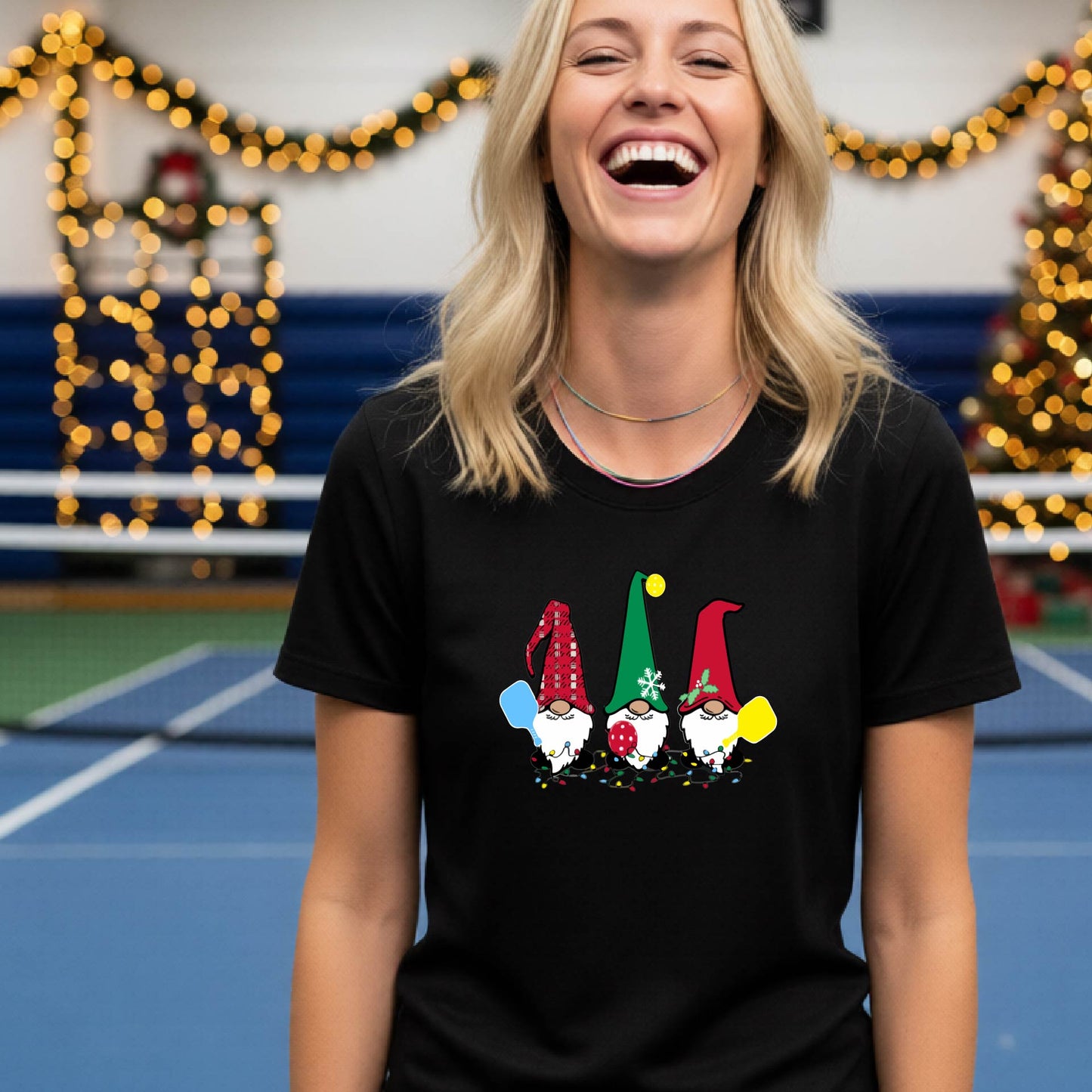 A smiling woman wearing a black graphic t-shirt with three cartoon Christmas pickleball gnomes, standing in a gymnasium decorated with holiday lights and a Christmas tree.