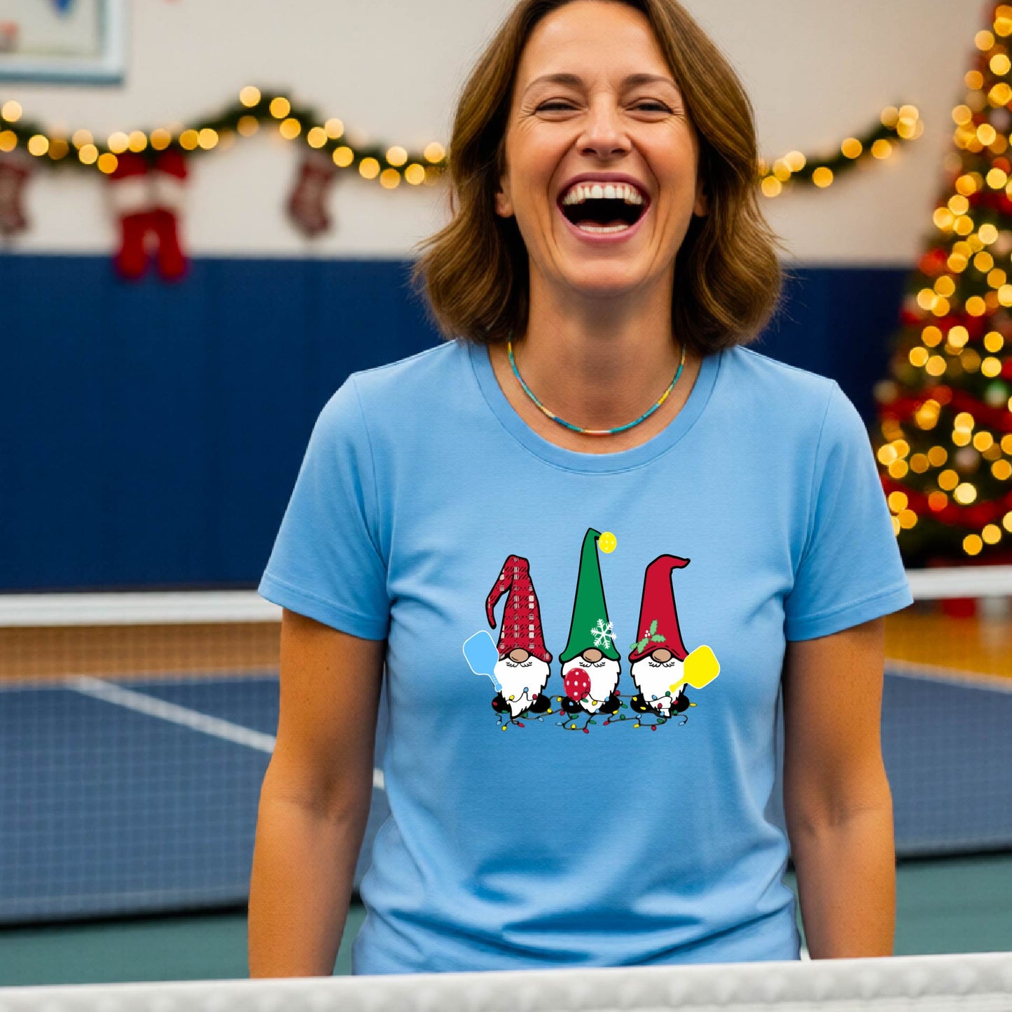 A smiling woman wearing a light blue graphic t-shirt with three cartoon Christmas pickleball gnomes, standing in a gymnasium decorated with holiday lights and a Christmas tree.