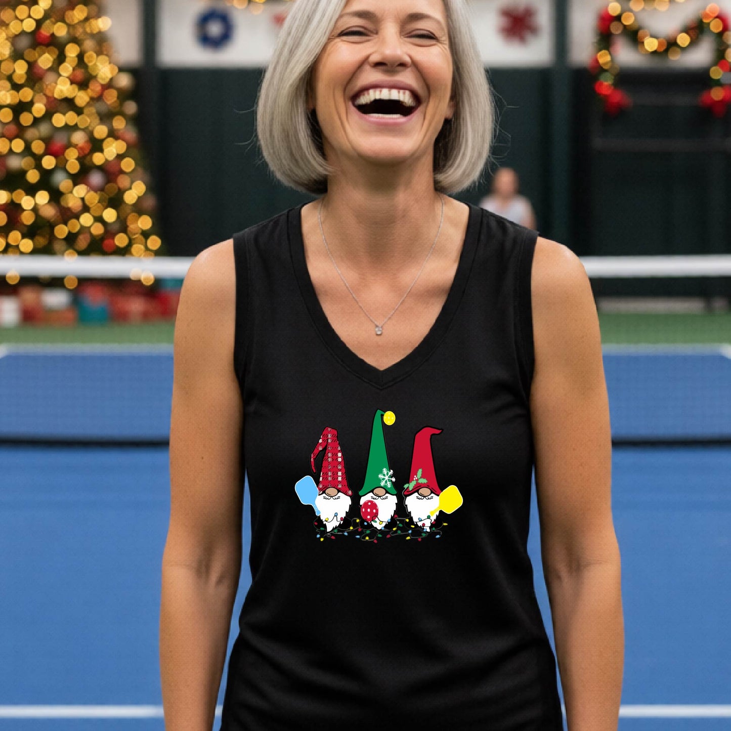 A woman laughing on a  pickleball court, wearing a black sleeveless shirt with three colorful gnome holding pickleball paddles graphic on the chest. The background features warm string lights and a decorated Christmas tree.