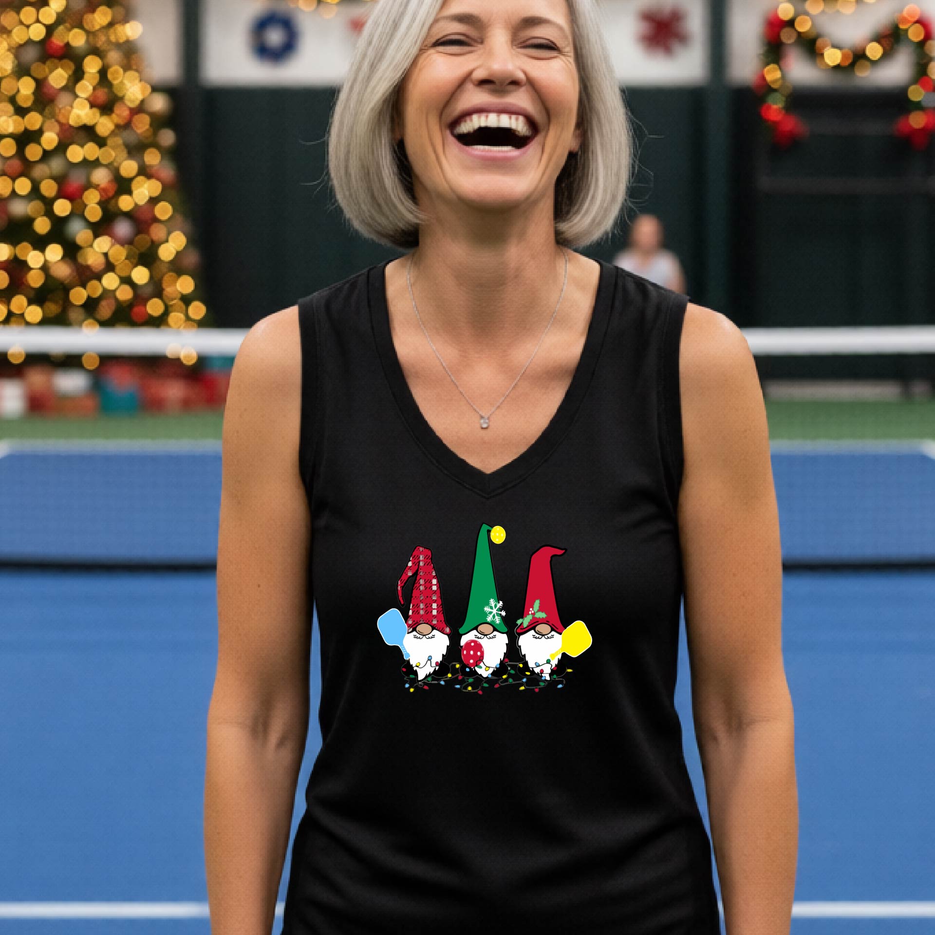 A woman laughing on a  pickleball court, wearing a black sleeveless shirt with three colorful gnome holding pickleball paddles graphic on the chest. The background features warm string lights and a decorated Christmas tree.