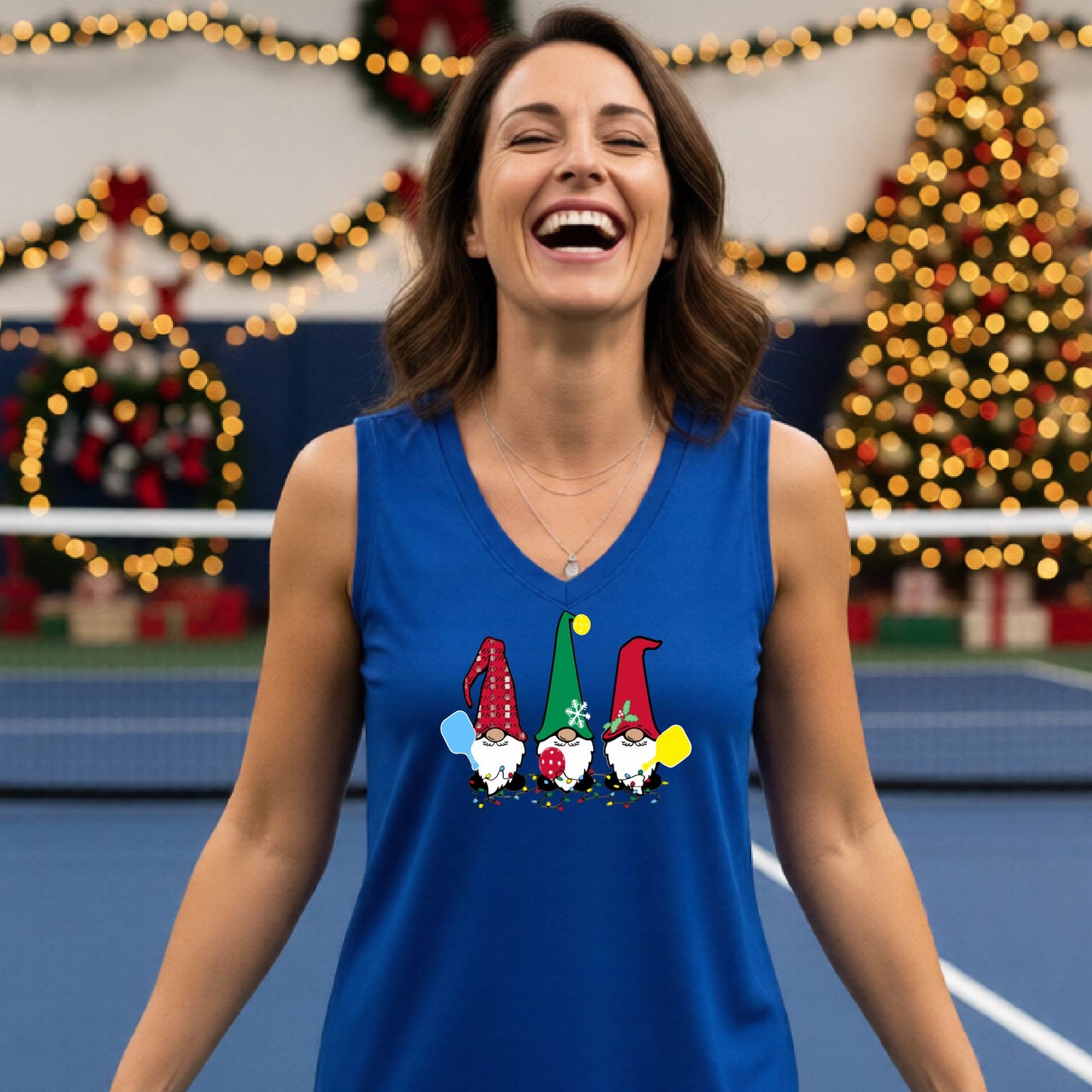 A woman laughing on a  pickleball court, wearing a royal blue sleeveless shirt with three colorful gnome holding pickleball paddles graphic on the chest. The background features warm string lights and a decorated Christmas tree.