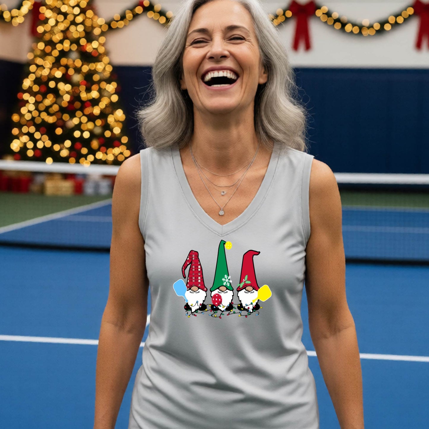A woman laughing on a  pickleball court, wearing a silver gray sleeveless shirt with three colorful gnome holding pickleball paddles graphic on the chest. The background features warm string lights and a decorated Christmas tree.