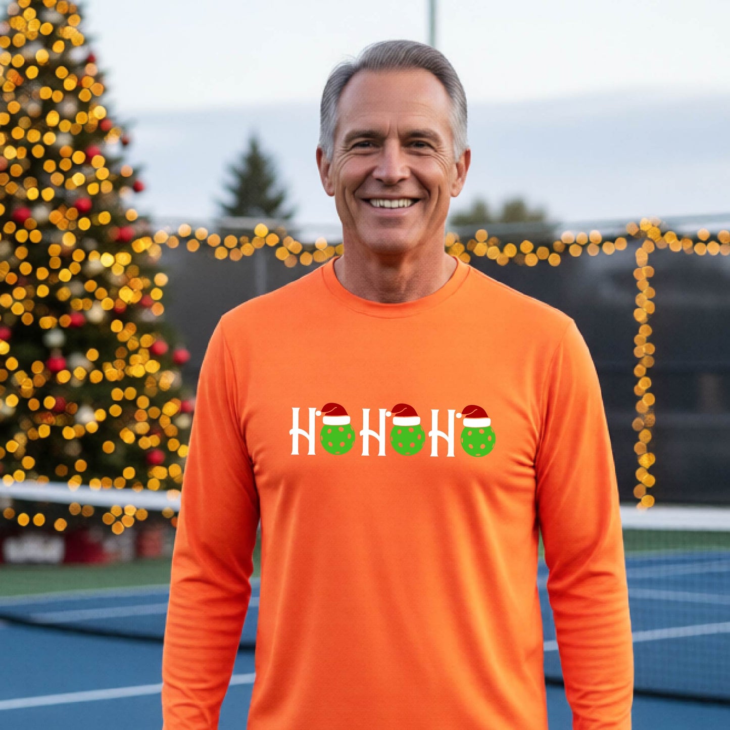 A smiling man wearing a bright orange long-sleeve athletic shirt with a pickleball Christmas-themed graphic of 'HOHOHO' with the O replaced  with green pickleballs wearing Santa hat stands in front of a blurred background featuring a lit Christmas tree and a pickleball court.