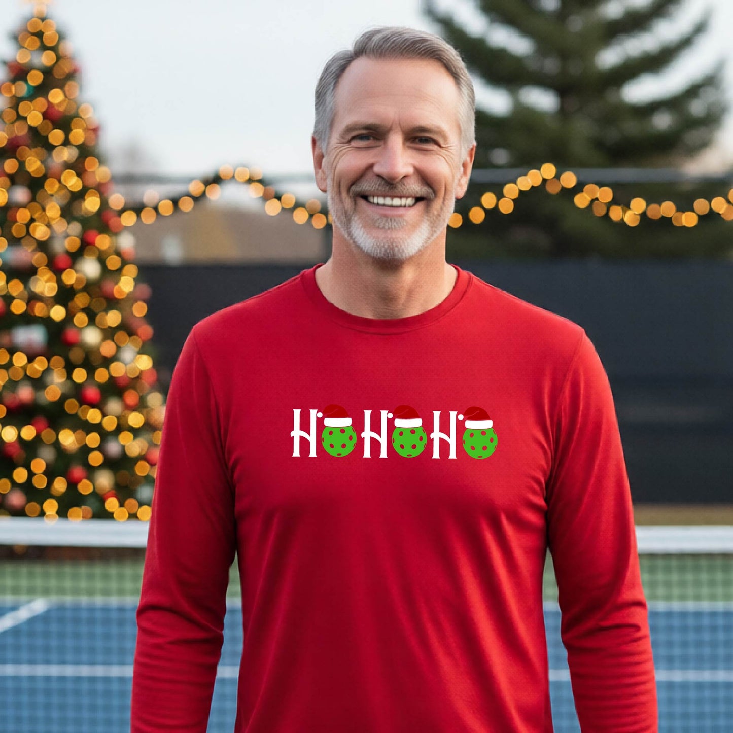 A smiling man wearing a red long-sleeve athletic shirt with a pickleball Christmas-themed graphic of 'HOHOHO' with the O replaced  with green pickleballs wearing Santa hat stands in front of a blurred background featuring a lit Christmas tree and a pickleball court.