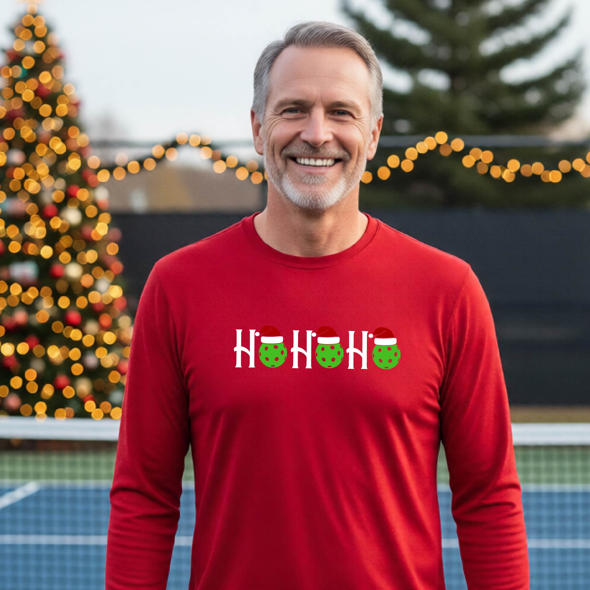 A smiling man wearing a red long-sleeve athletic shirt with a pickleball Christmas-themed graphic of 'HOHOHO' with the O replaced  with green pickleballs wearing Santa hat stands in front of a blurred background featuring a lit Christmas tree and a pickleball court.