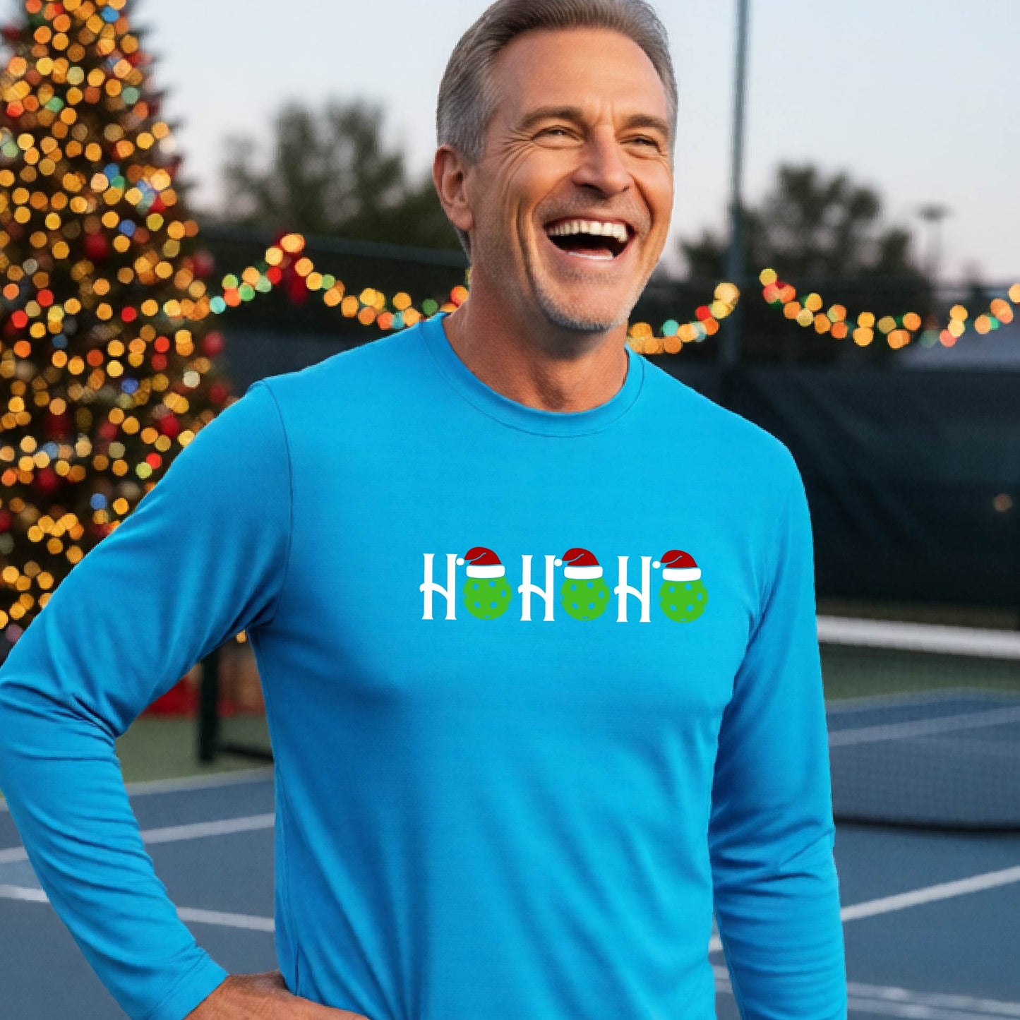 A smiling man wearing a bright blue long-sleeve athletic shirt with a pickleball Christmas-themed graphic of 'HOHOHO' with the O replaced  with green pickleballs wearing Santa hat stands in front of a blurred background featuring a lit Christmas tree and a pickleball court.