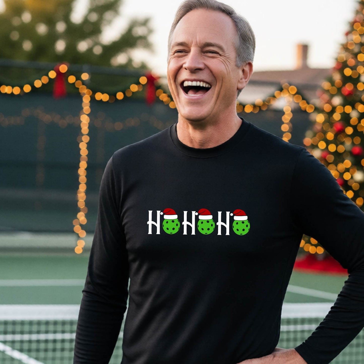 A smiling man wearing a black long-sleeve athletic shirt with a pickleball Christmas-themed graphic of 'HOHOHO' with the O replaced  with green pickleballs wearing Santa hat stands in front of a blurred background featuring a lit Christmas tree and a pickleball court.