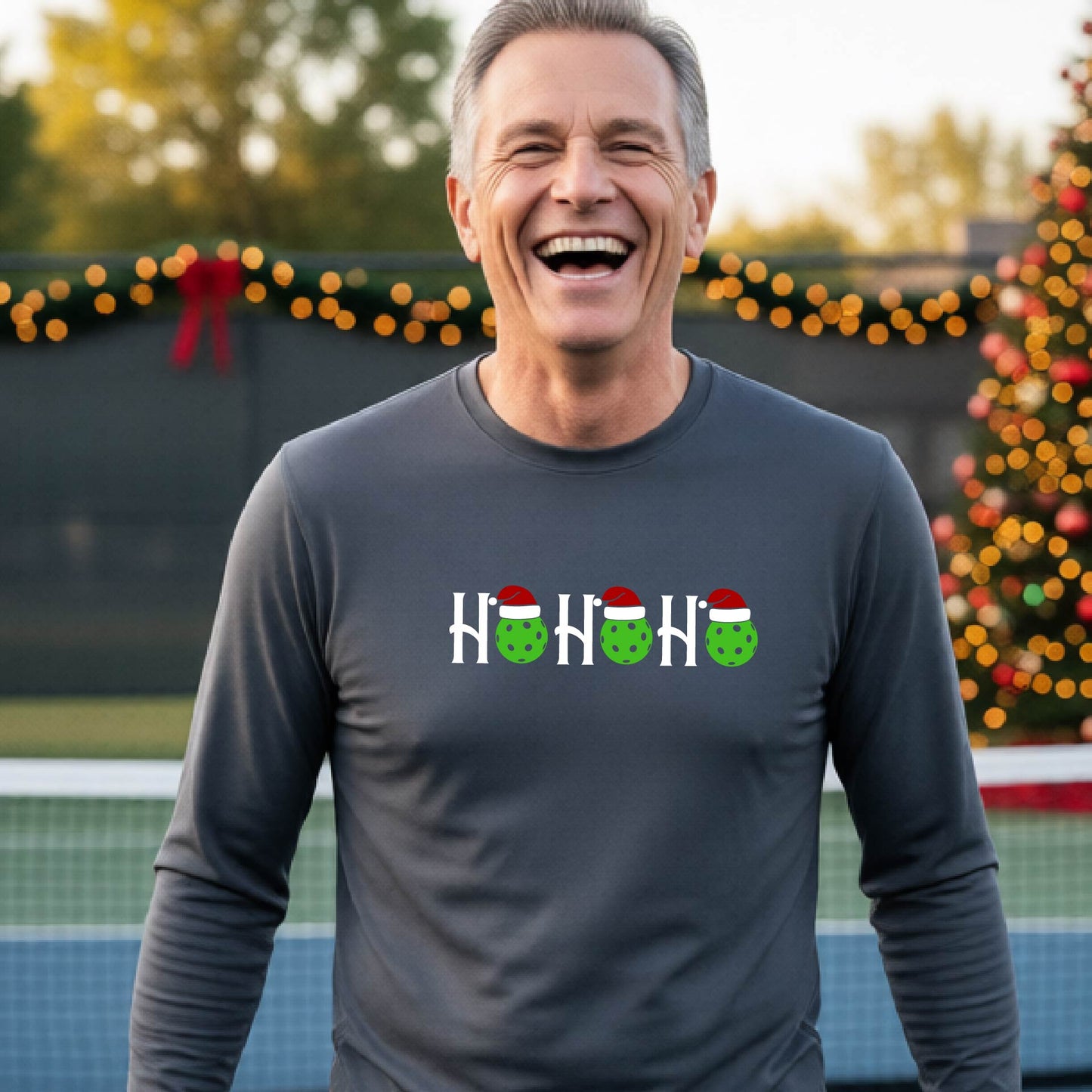 A smiling man wearing a gray long-sleeve athletic shirt with a pickleball Christmas-themed graphic of 'HOHOHO' with the O replaced  with green pickleballs wearing Santa hat stands in front of a blurred background featuring a lit Christmas tree and a pickleball court.