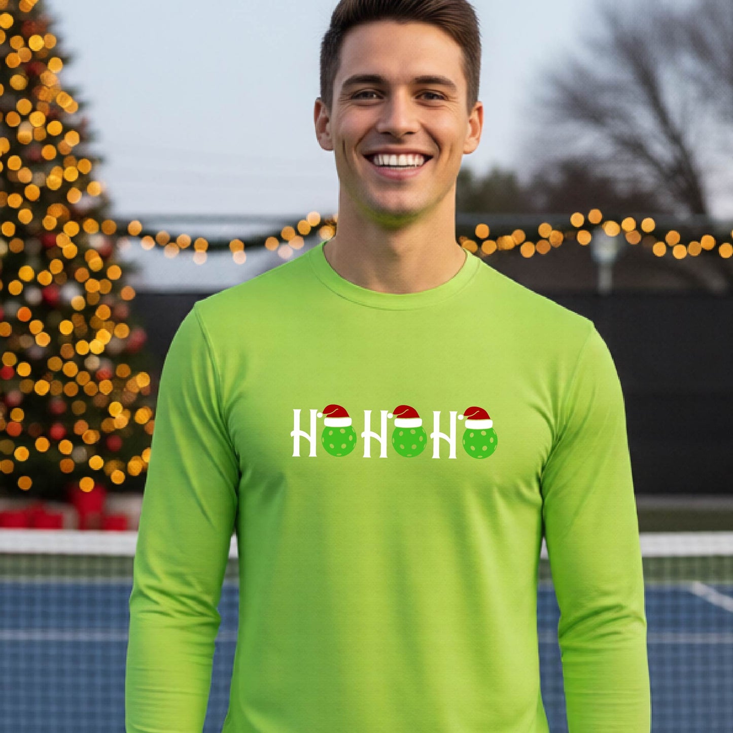 A smiling man wearing a lime green long-sleeve athletic shirt with a pickleball Christmas-themed graphic of 'HOHOHO' with the O replaced  with green pickleballs wearing Santa hat stands in front of a blurred background featuring a lit Christmas tree and a pickleball court.