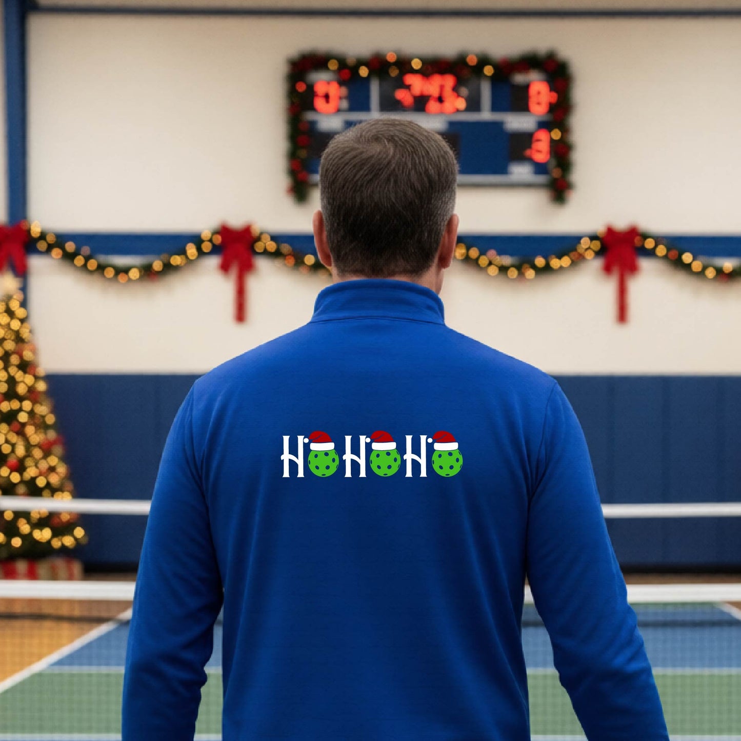A man viewed from behind in an indoor pickleball court, wearing a royal blue long-sleeve quarter-zip shirt with a Christmas-themed 'HO HO HO' pickleball wearing a Santa hat graphic on the back. 