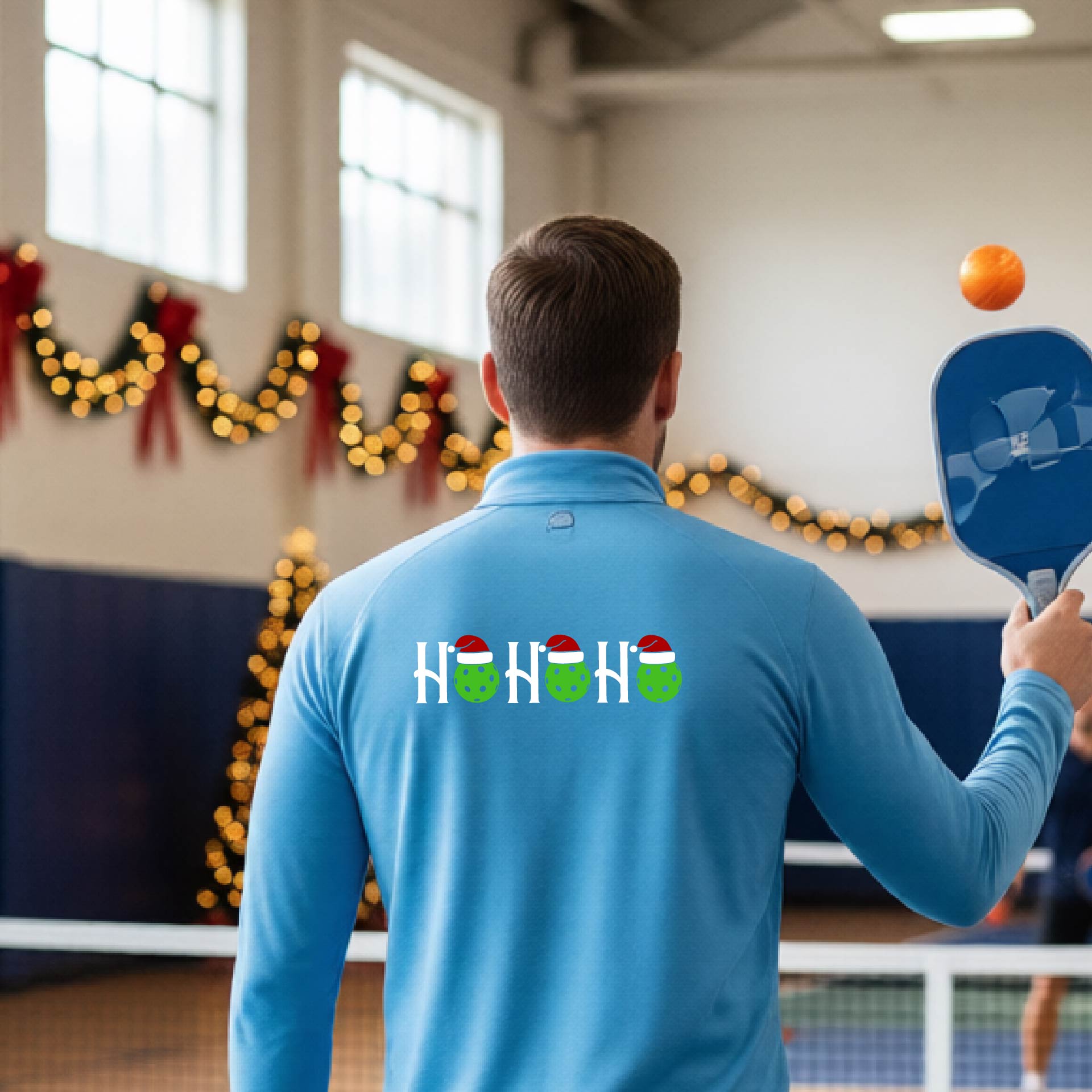 A man viewed from behind in an indoor pickleball court, wearing a light blue long-sleeve quarter-zip shirt with a Christmas-themed 'HO HO HO' pickleball wearing a Santa hat graphic on the back. 