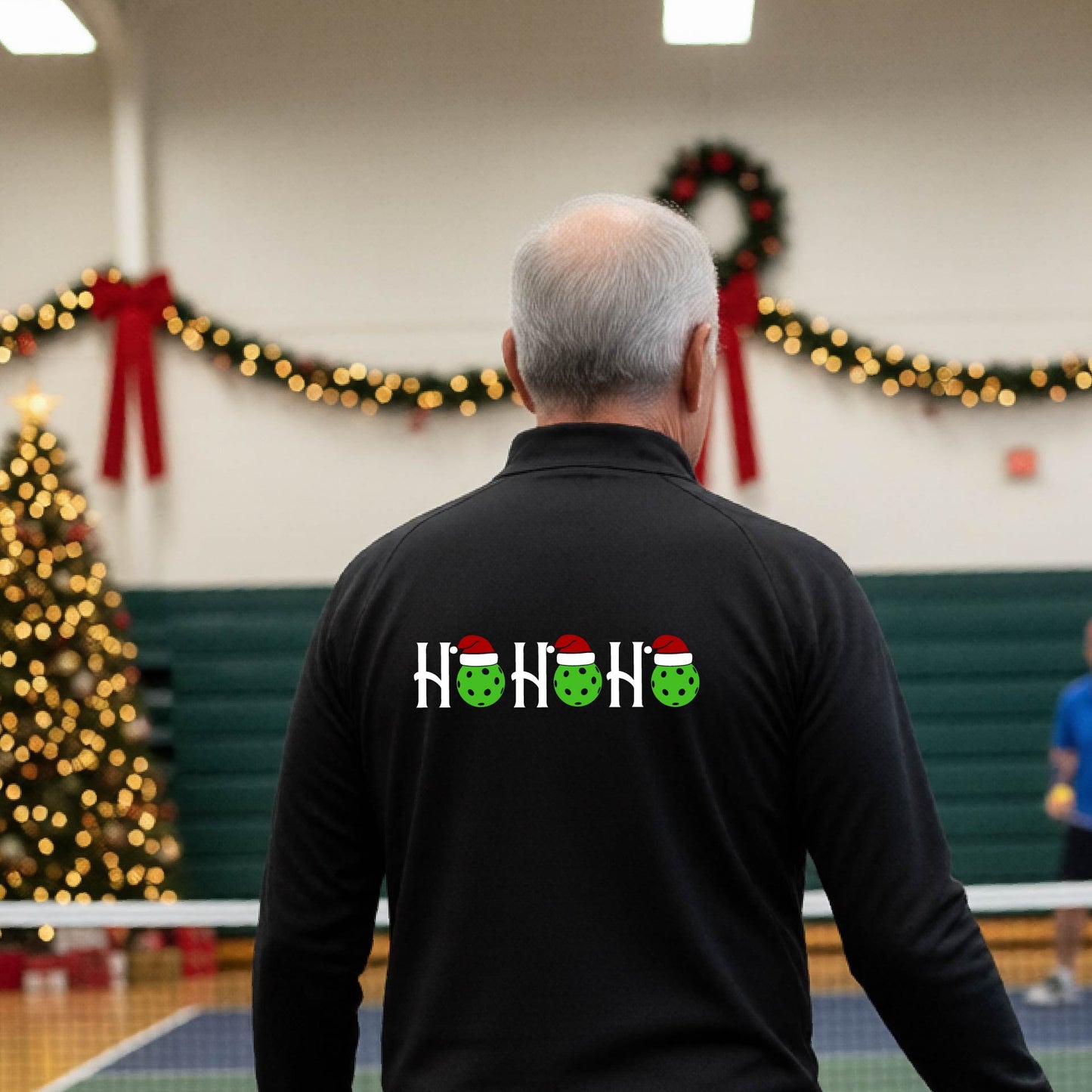 A man viewed from behind in an indoor pickleball court, wearing a black long-sleeve quarter-zip shirt with a Christmas-themed 'HO HO HO' pickleball wearing a Santa hat graphic on the back. 