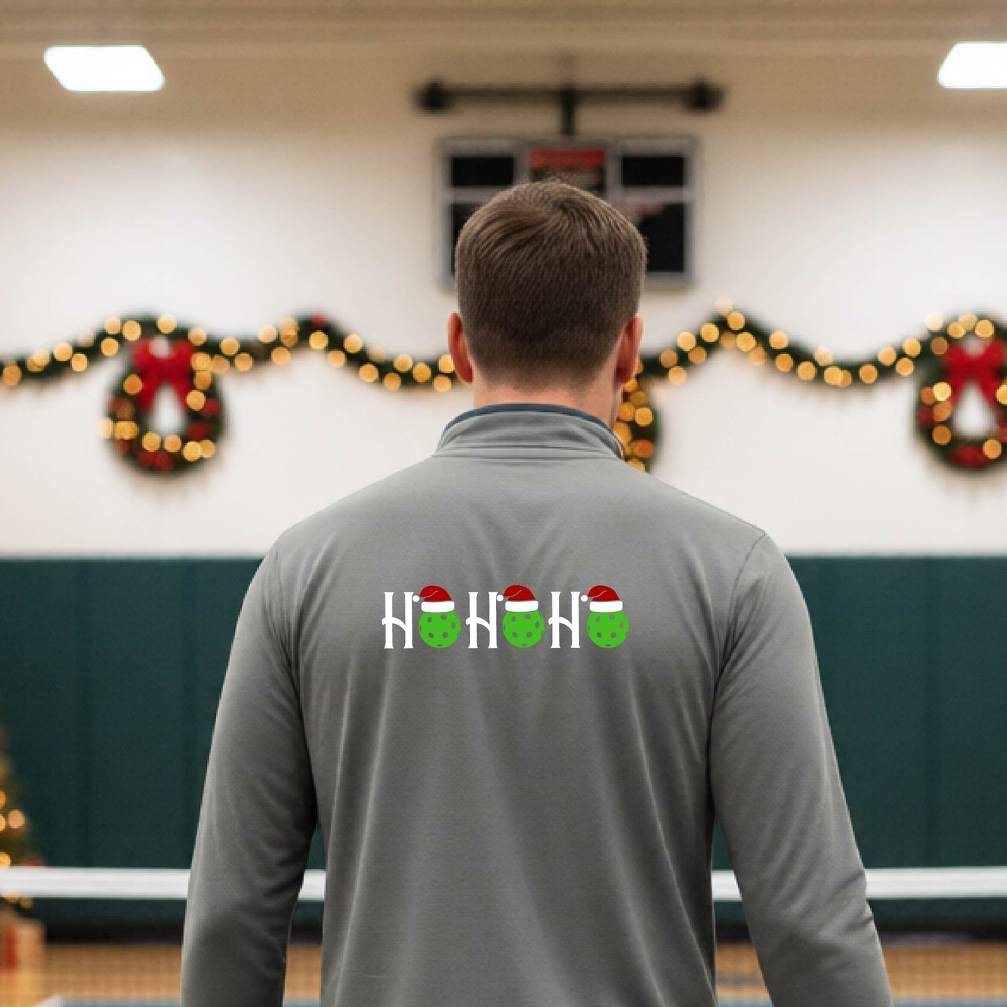 A man viewed from behind in an indoor pickleball court, wearing a light grey long-sleeve quarter-zip shirt with a Christmas-themed 'HO HO HO' pickleball wearing a Santa hat graphic on the back. 