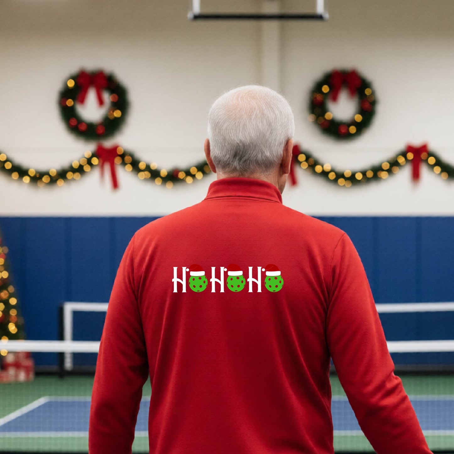 A man viewed from behind in an indoor pickleball court, wearing a red long-sleeve quarter-zip shirt with a Christmas-themed 'HO HO HO' pickleball wearing a Santa hat graphic on the back. 