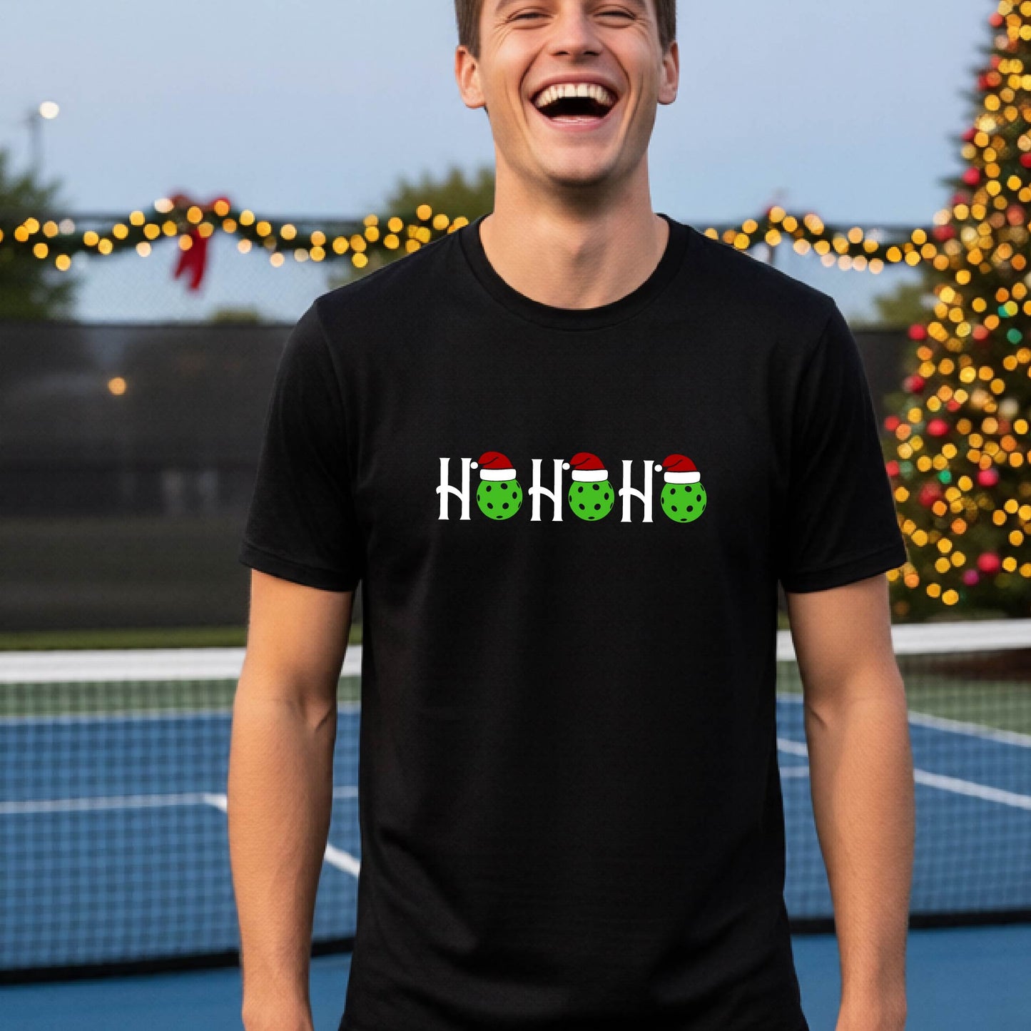 A man wearing a black short-sleeve t-shirt with a 'HO HO HO' graphic made of Christmas pickleballs wearing red Santa hats, set against a blurred background of a pickleball court and a decorated Christmas tree.